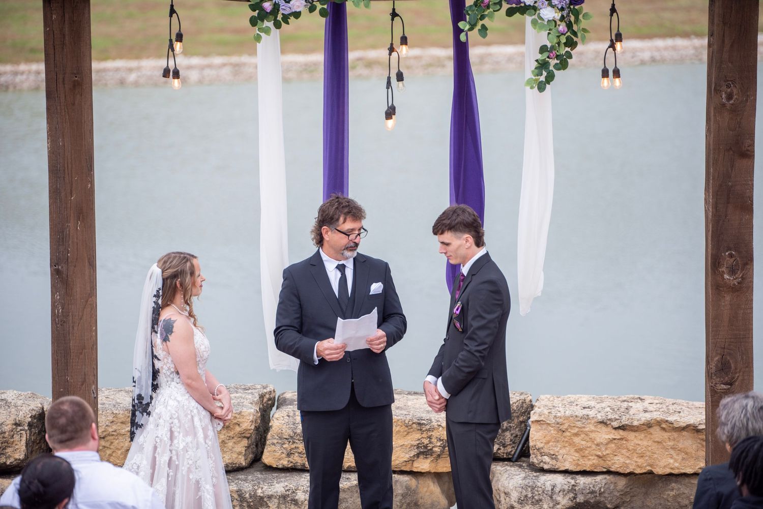 Outdoor wedding ceremony taking place under a wooden pergola by the waterfront.