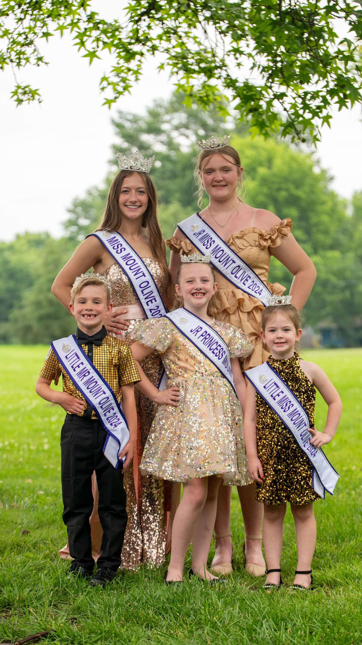 Group photo of pageant contestants wearing sparkly dresses and sashes posing together on green grass.