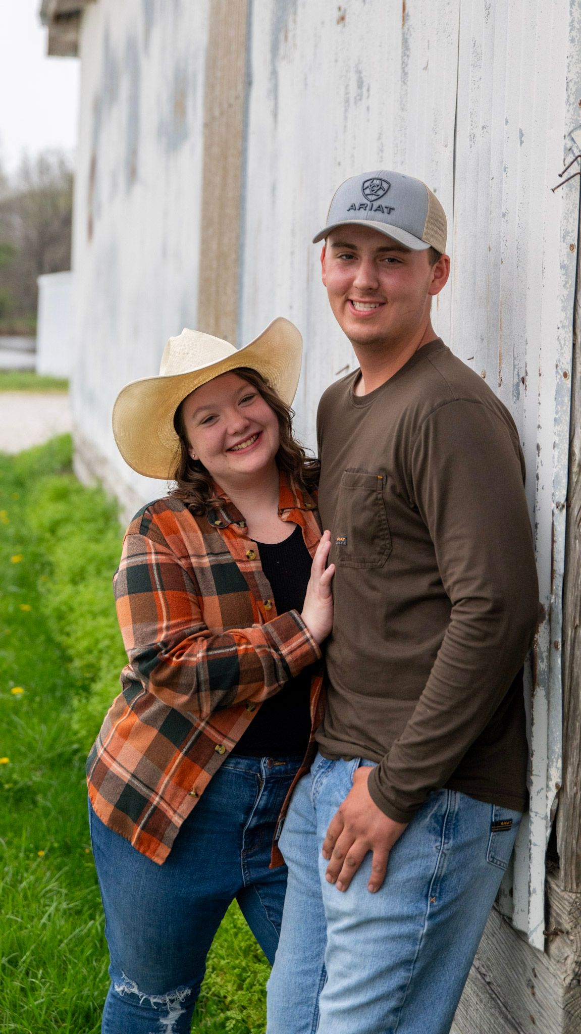 Couple in casual attire poses against weathered white barn wall.