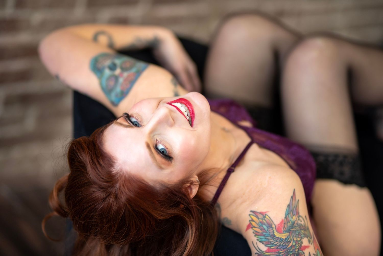 Woman with red hair and tattoos poses on floor in black lingerie for boudoir photography session.