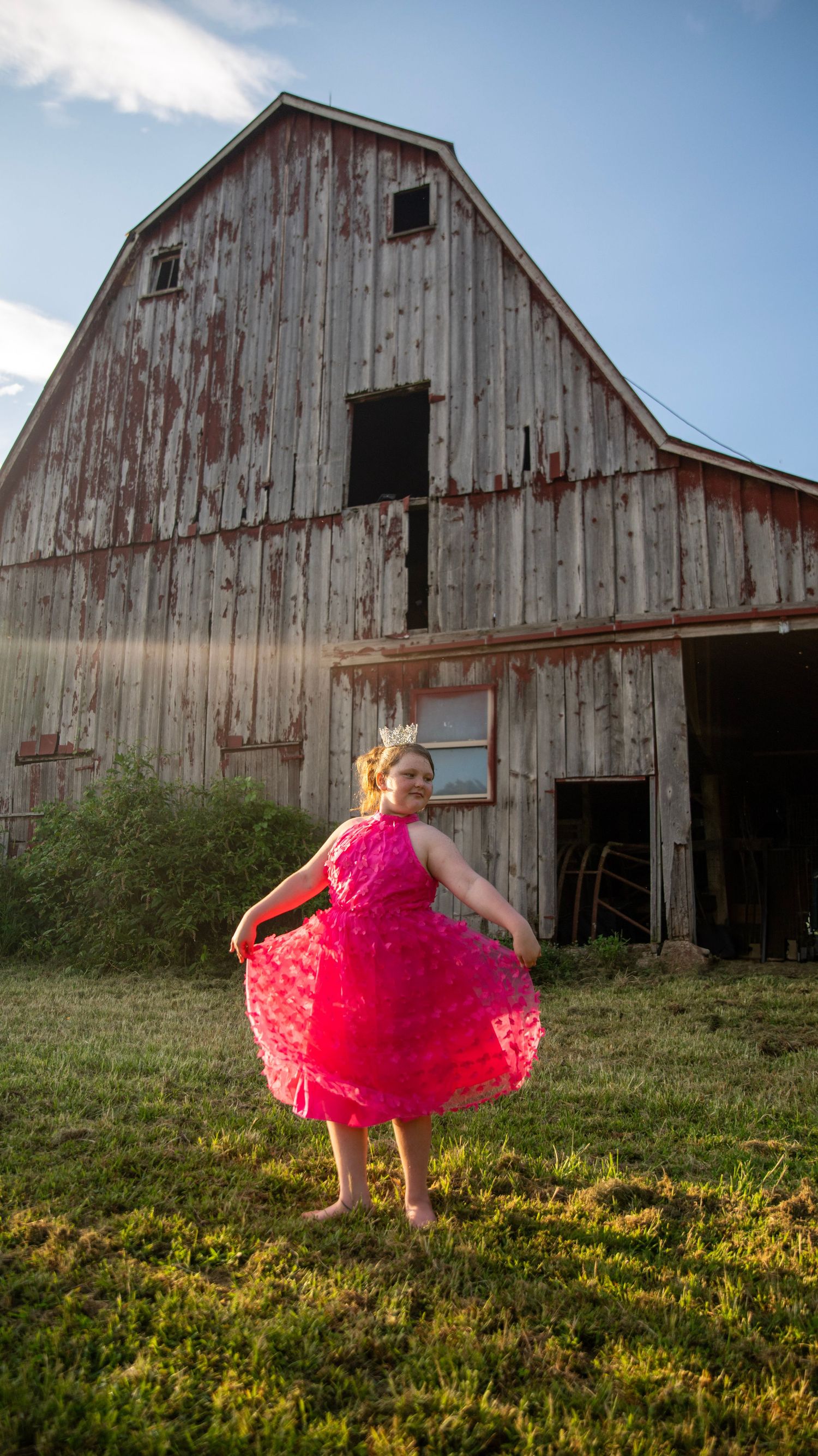 Person in bright pink dress twirls in front of rustic barn during golden hour.