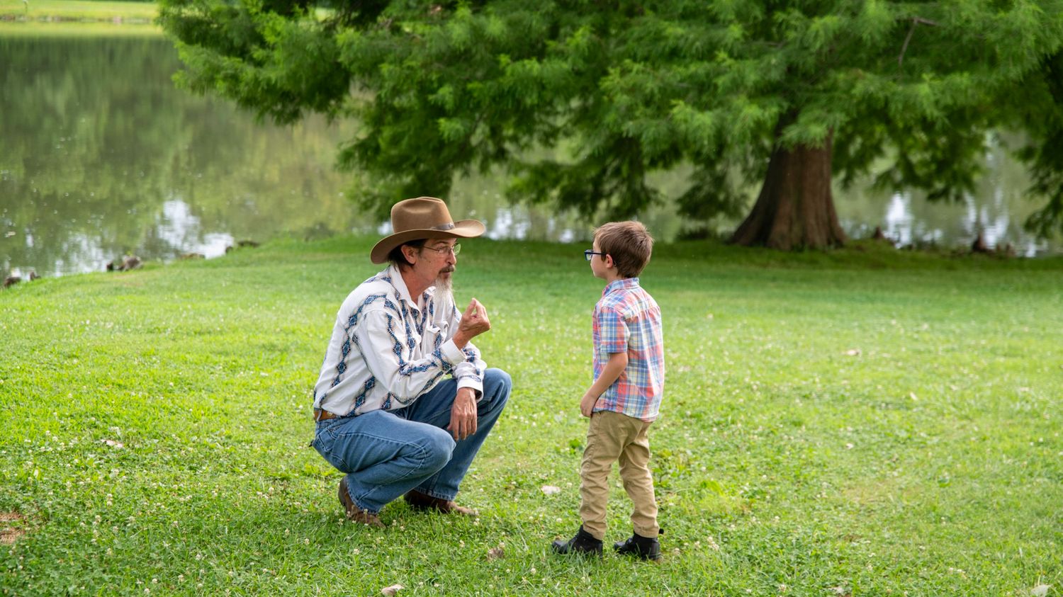 Two figures share a moment on green grass with trees and pond in background.