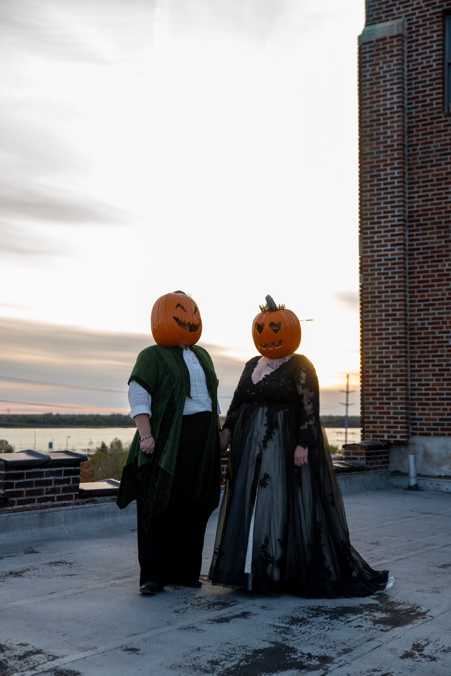 Two people in gothic attire with pumpkin heads standing on rooftop at sunset.