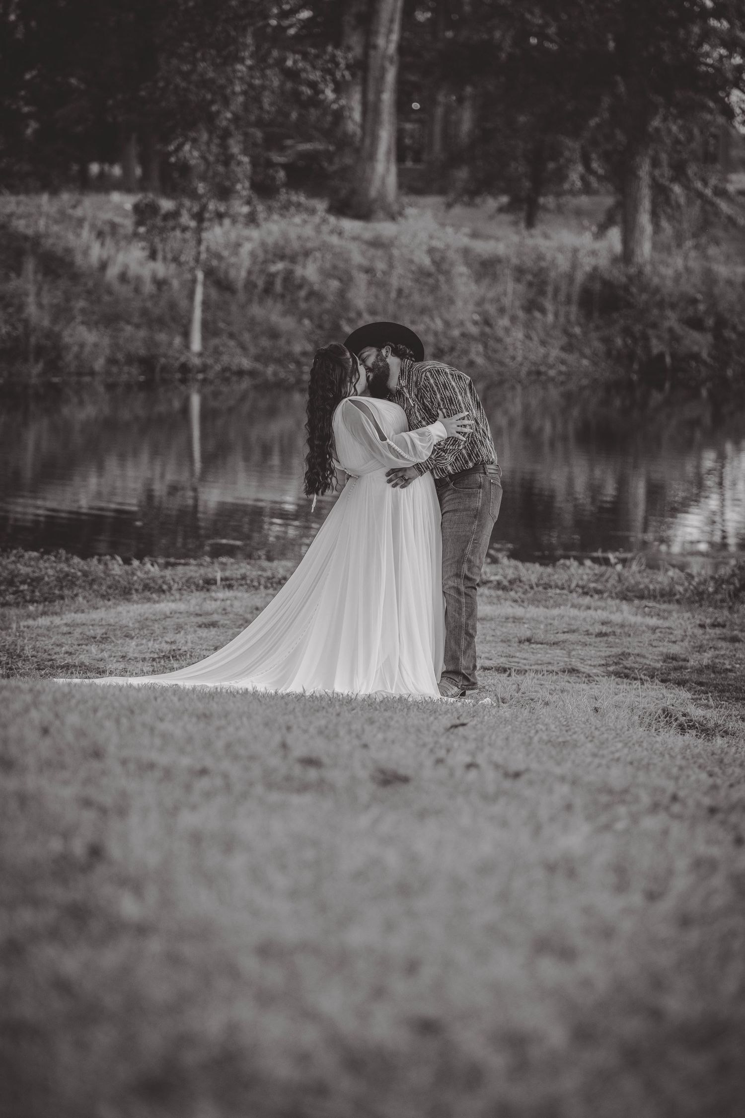 Black and white wedding photo of a couple by a serene lake with trees in the background.