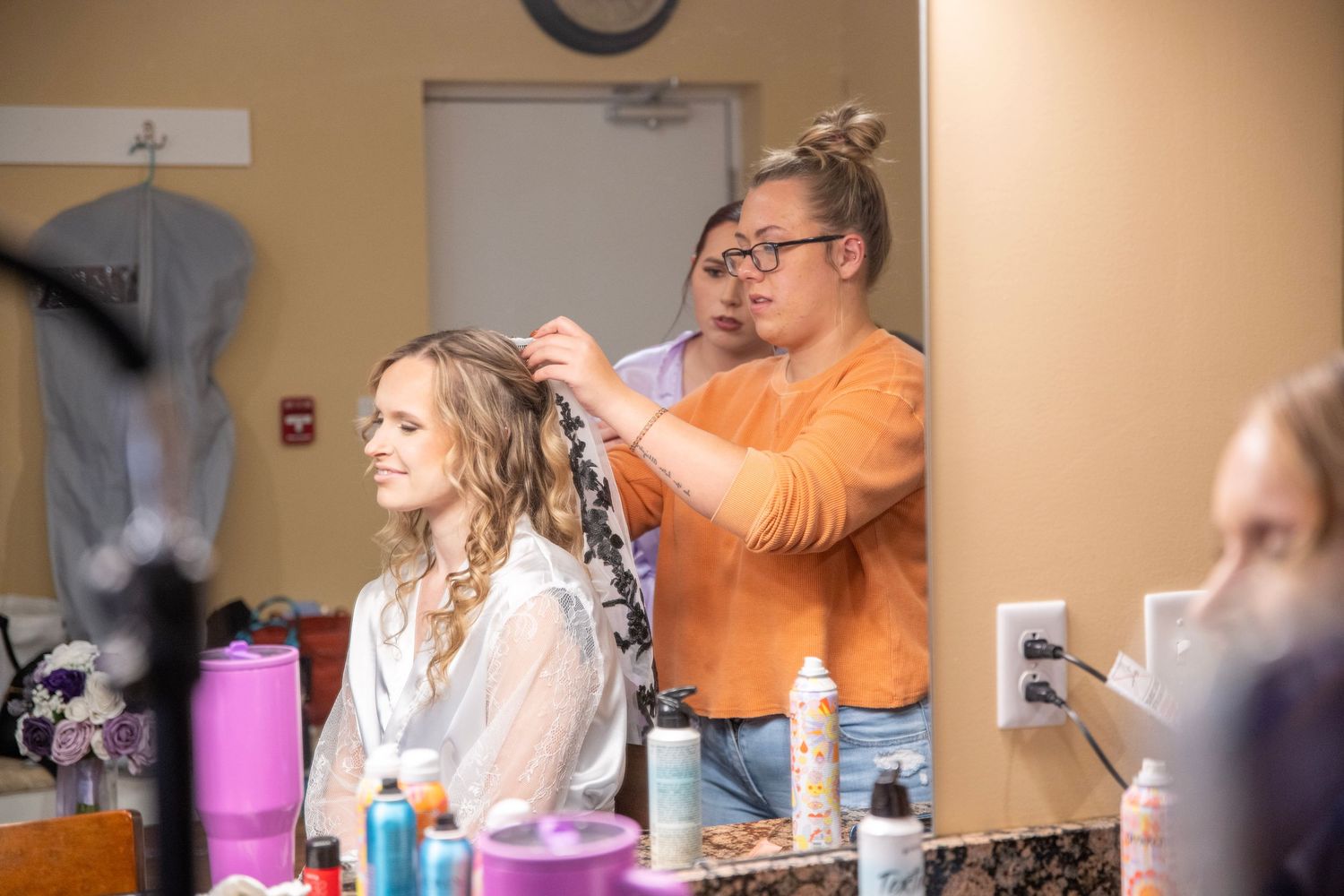 Stylists work on hair and makeup preparations in a warm-lit getting ready room.