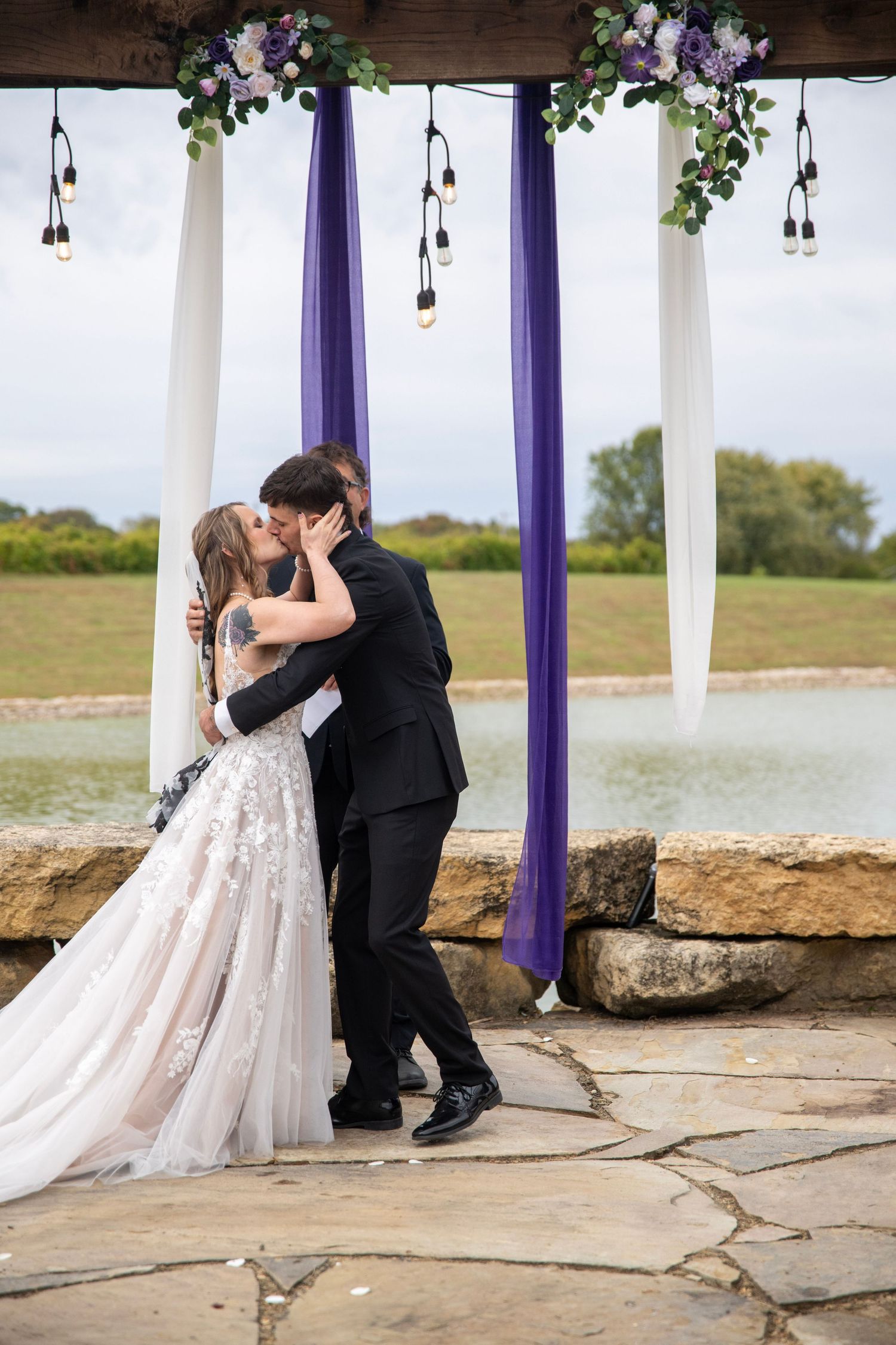 Romantic first kiss as newlyweds under pergola with purple and white draping.