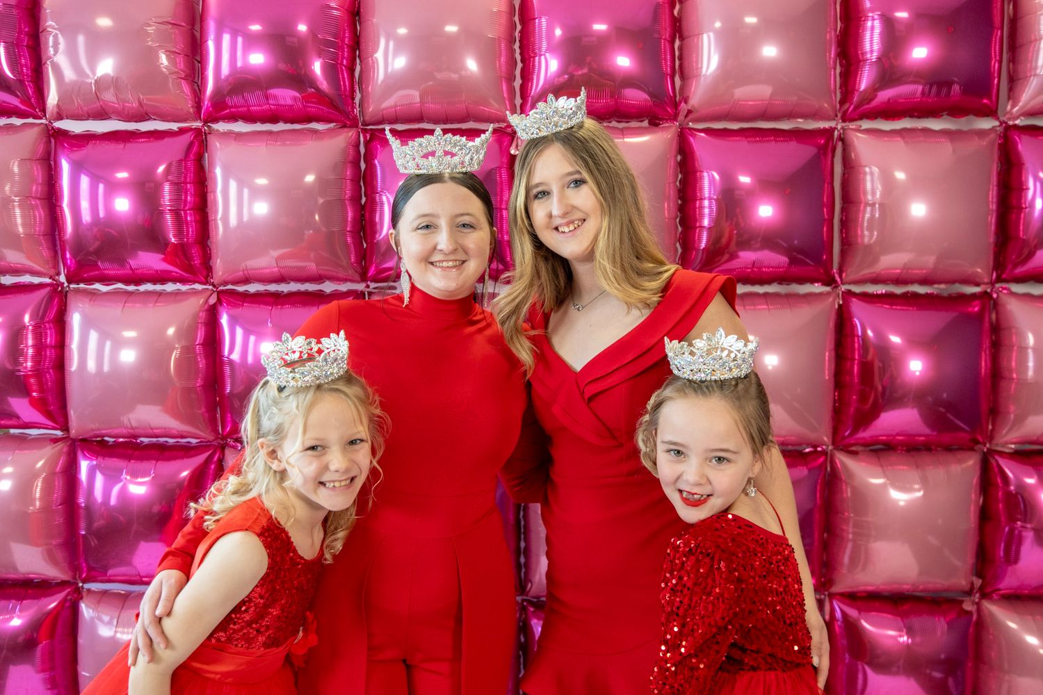 Group in matching red outfits pose together against pink balloon wall.