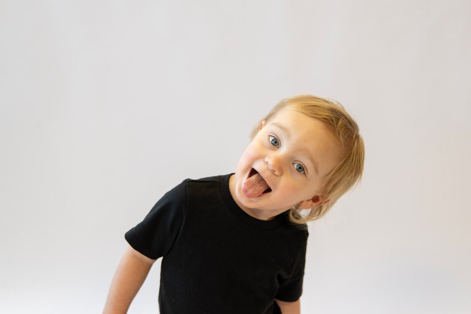 Energetic toddler in black t-shirt making silly faces against white backdrop.