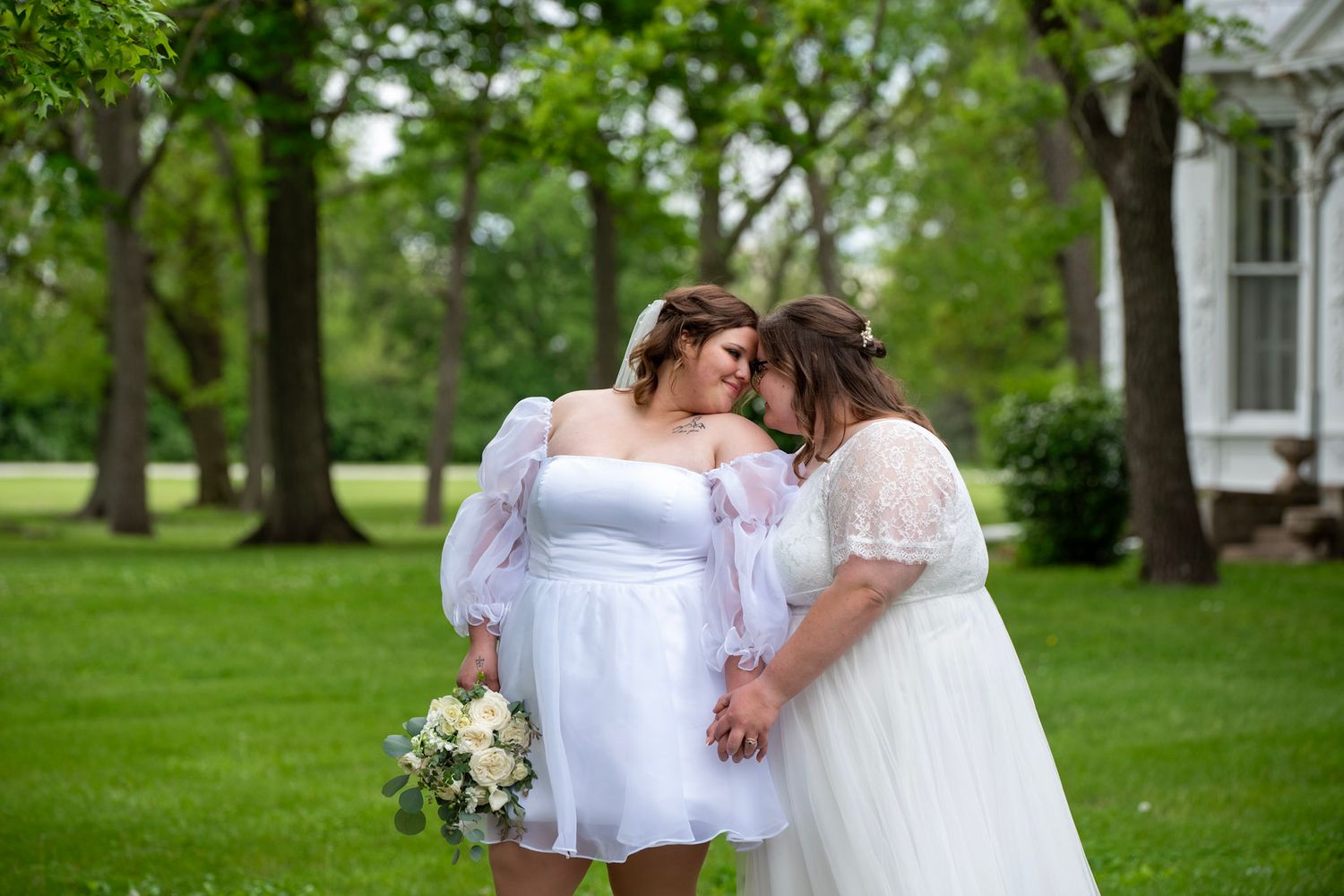 Couple in white outfits embrace in a green outdoor garden setting.