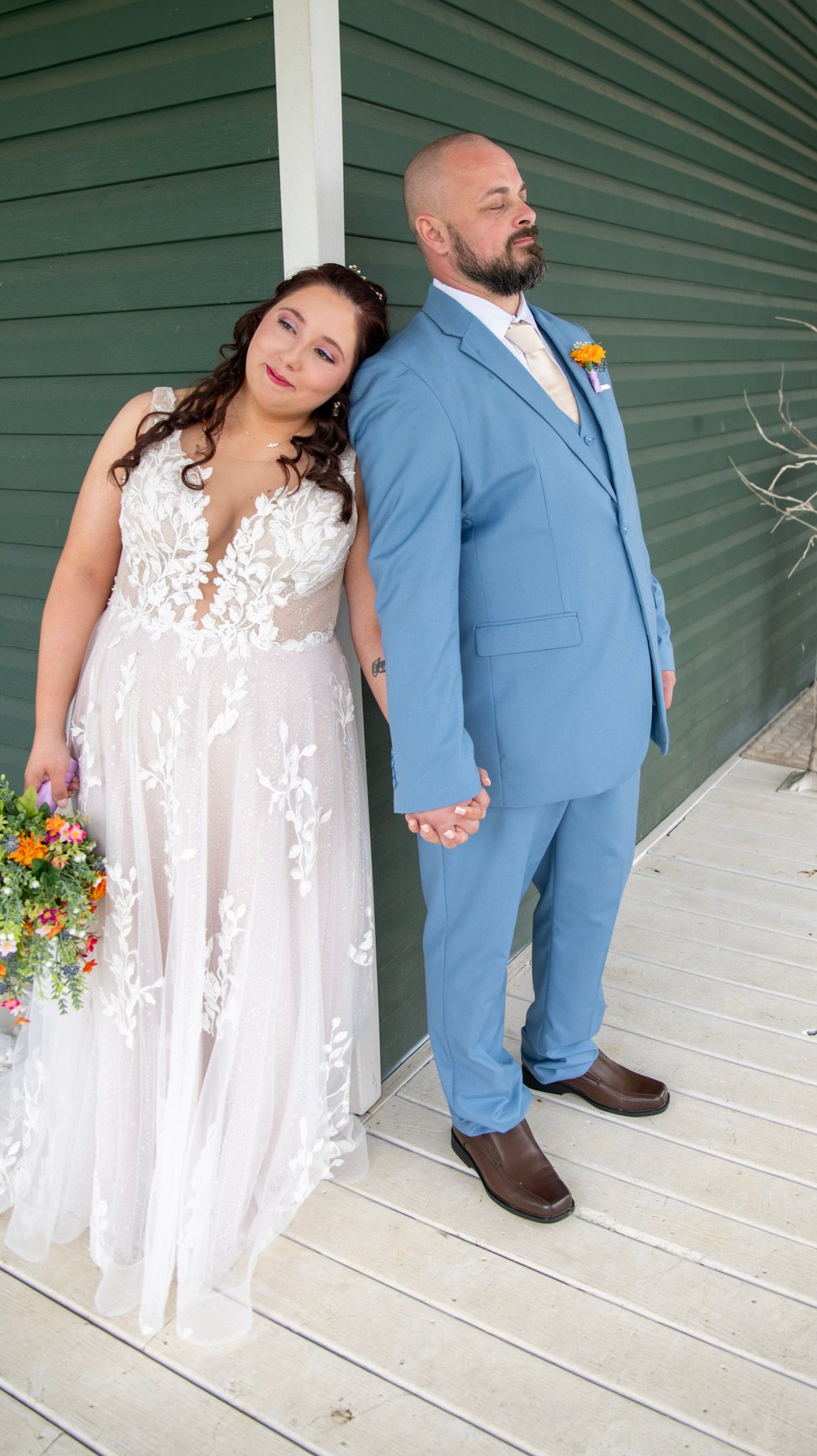 Couple in blue suit and white lace dress pose romantically on a green porch.