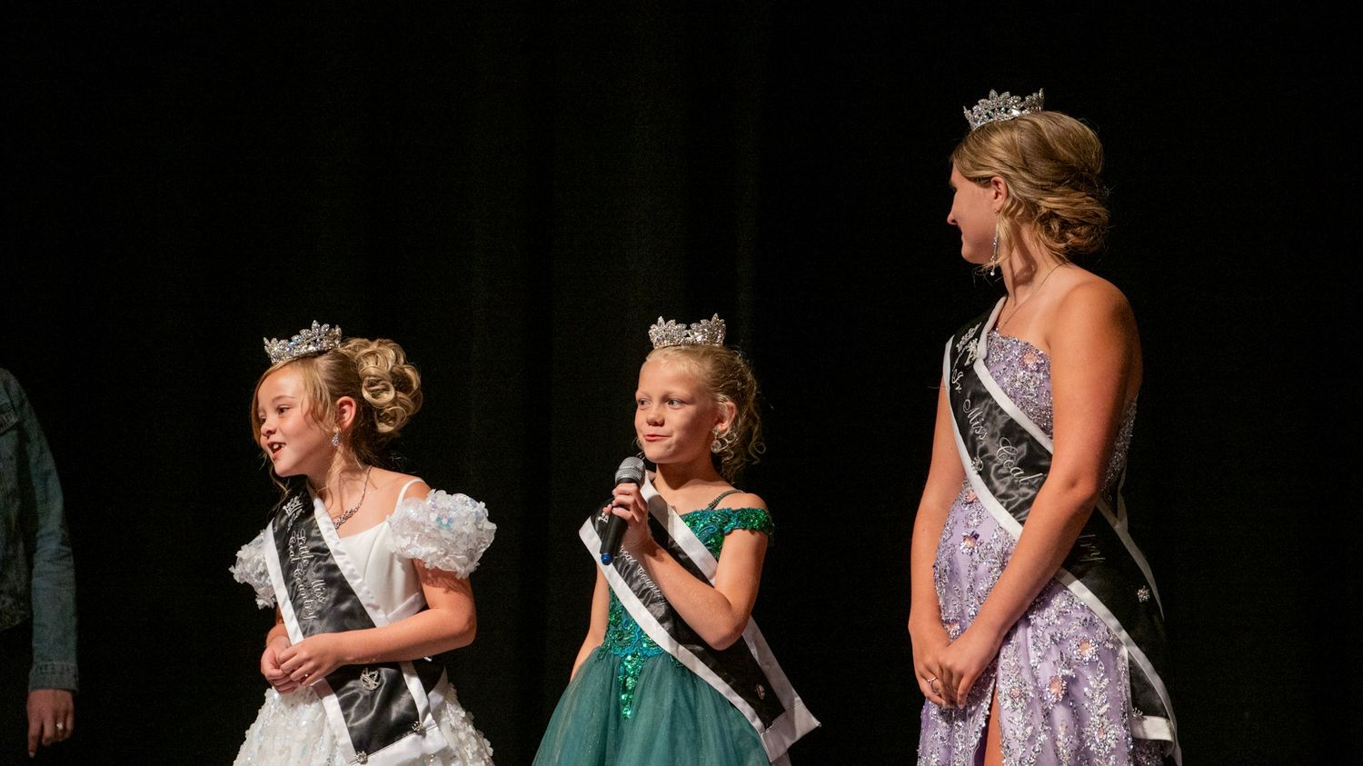 Pageant contestants in formal gowns stand together on stage wearing sashes.