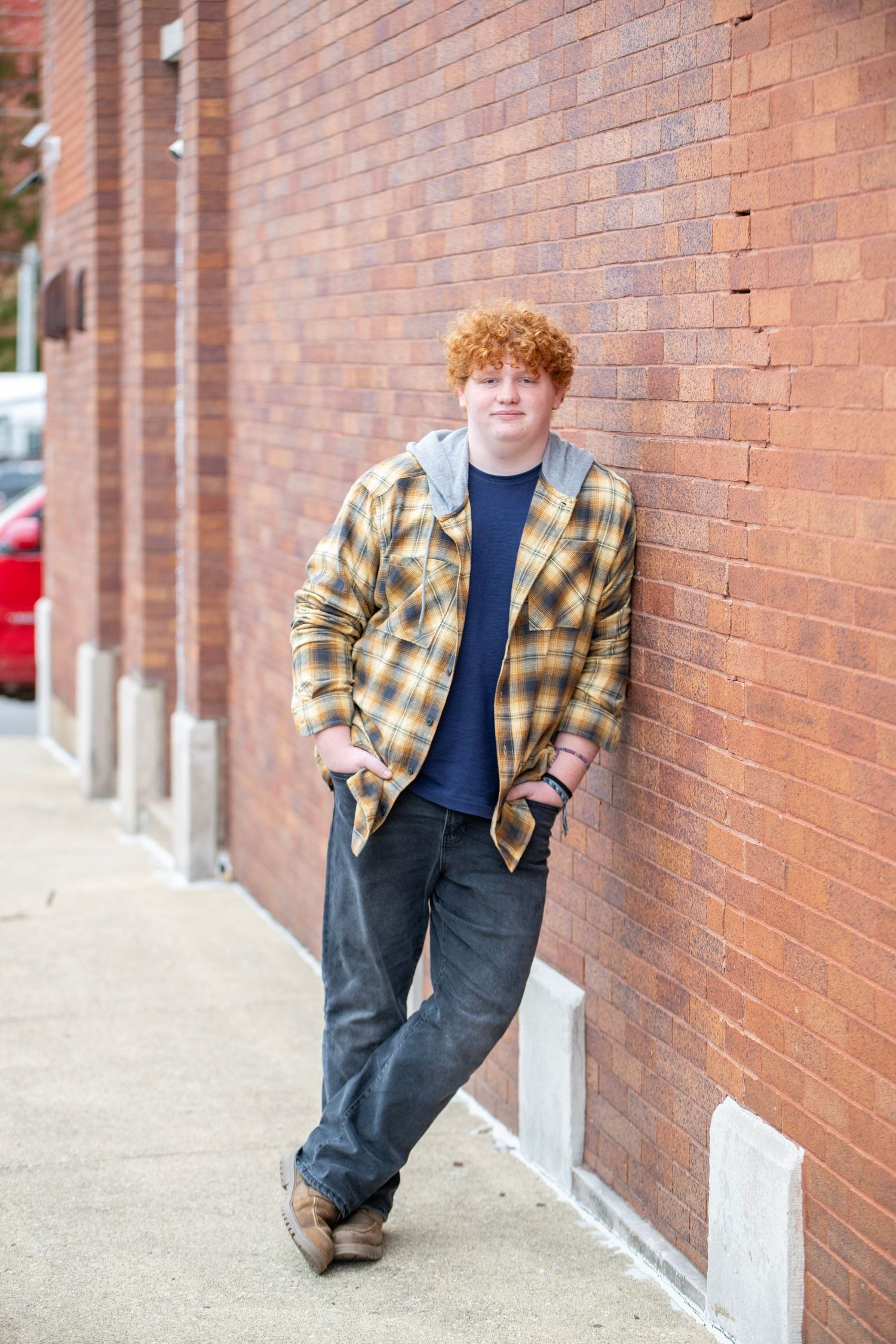 Portrait against brick wall wearing yellow plaid flannel shirt and dark pants in casual pose.