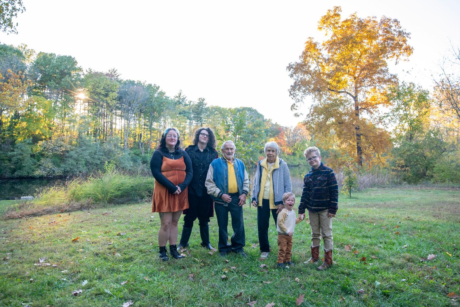 Multi-generational family gathering outdoors with fall colors and trees in background.