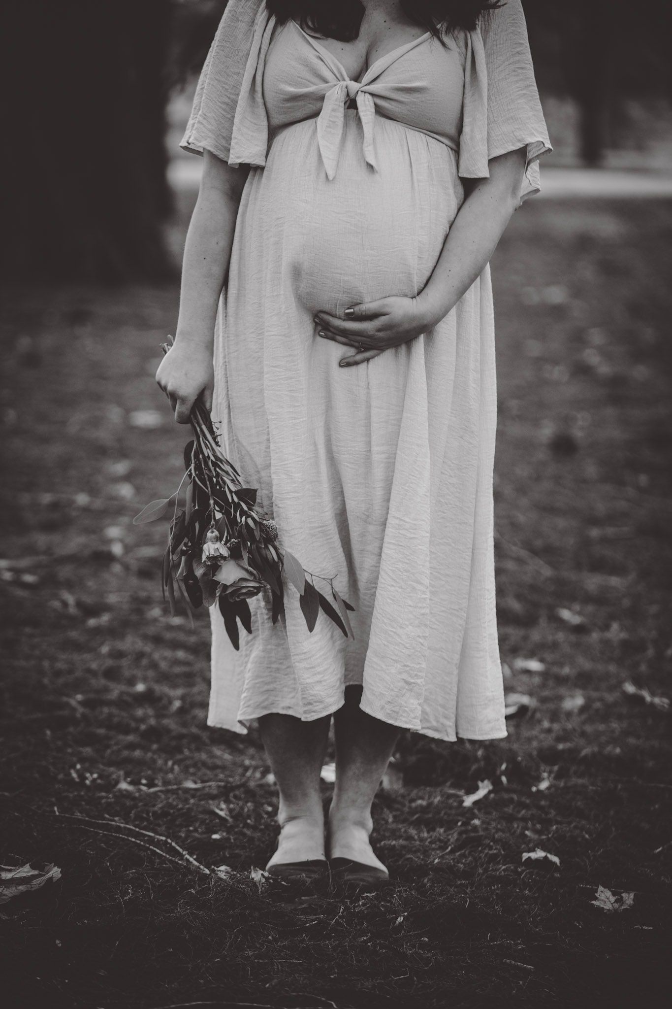 A black and white photo of a pregnant figure in a flowing dress holding dried flowers.