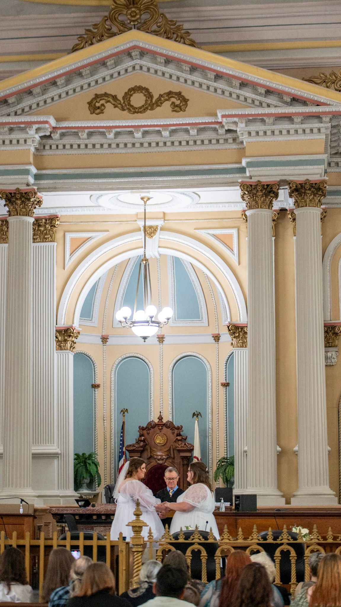 Ornate historic courthouse interior with white columns and blue walls during a wedding ceremony.