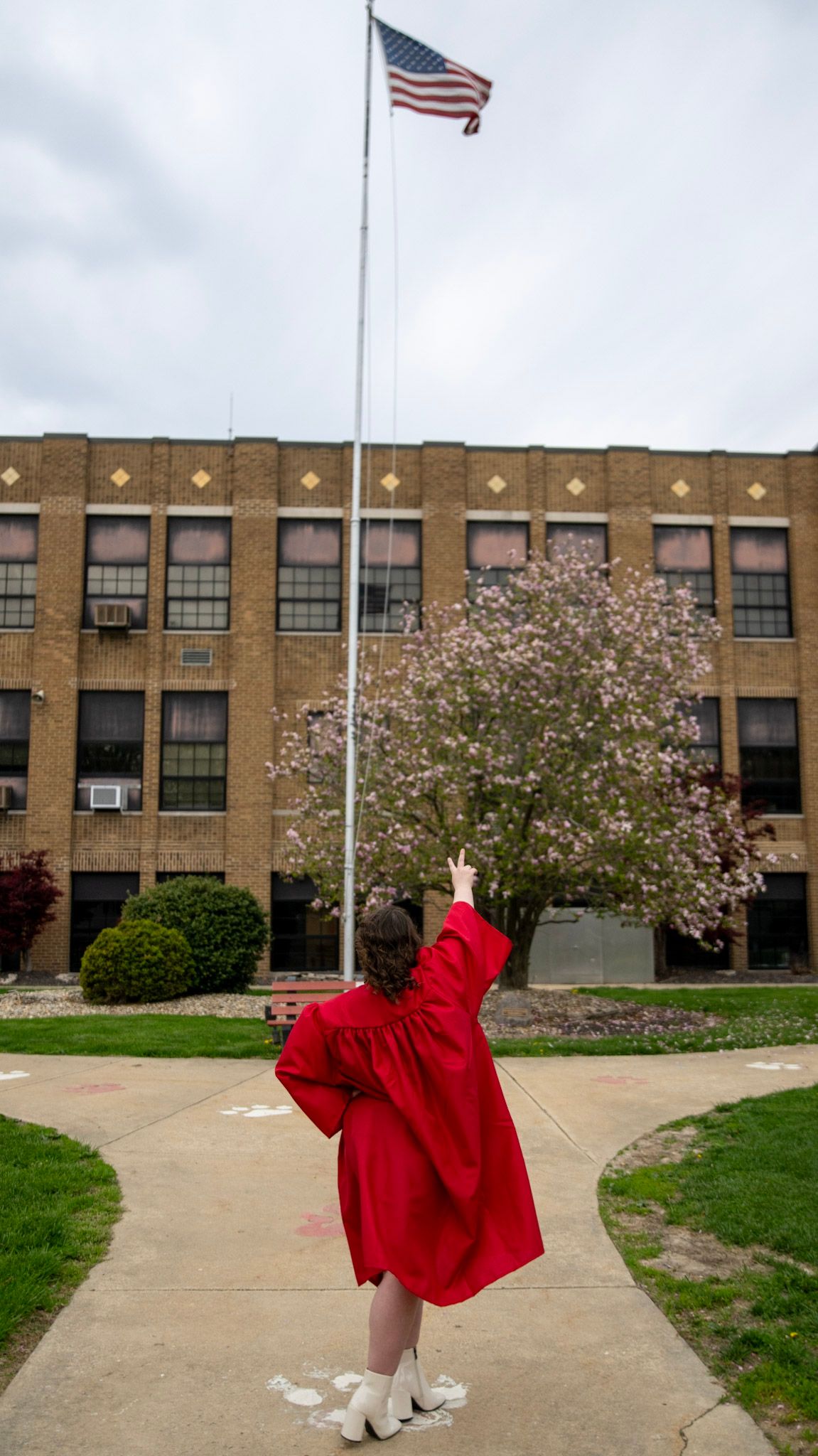 Graduate in red gown leaps joyfully in front of school building with American flag.