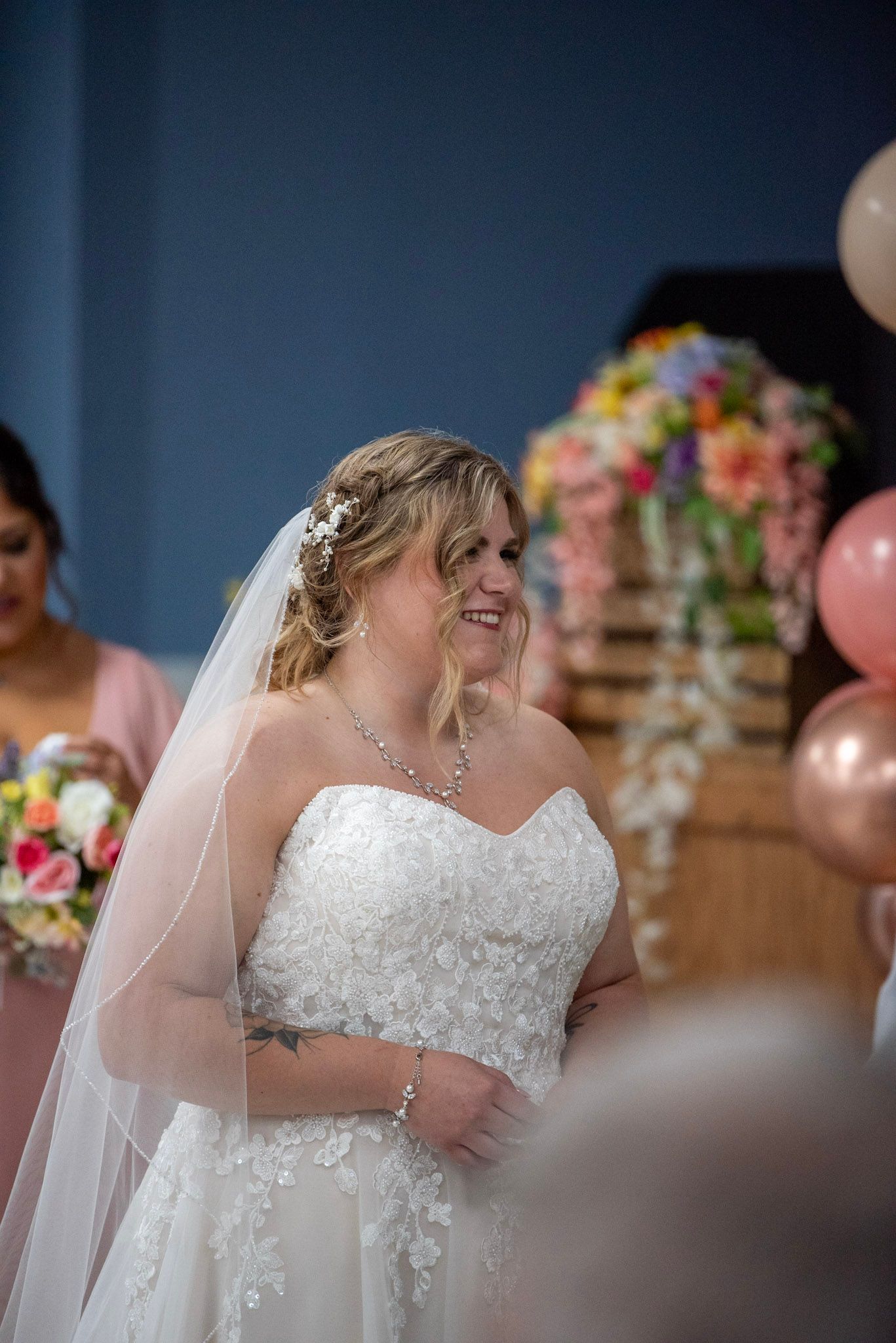 A bride in a lace wedding gown smiles during her ceremony with floral and balloon decorations.