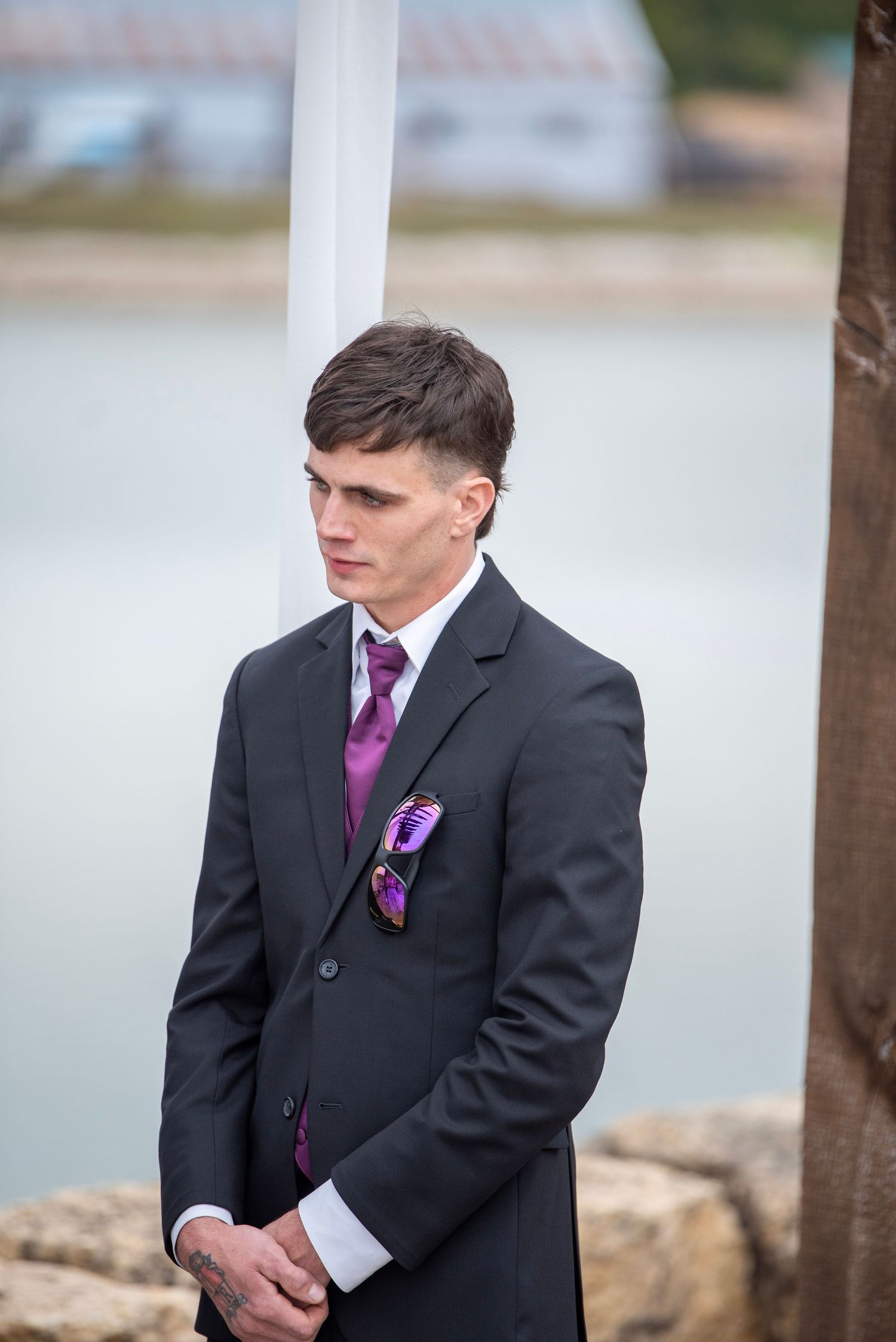 Person in black suit with purple accents standing at outdoor ceremony.