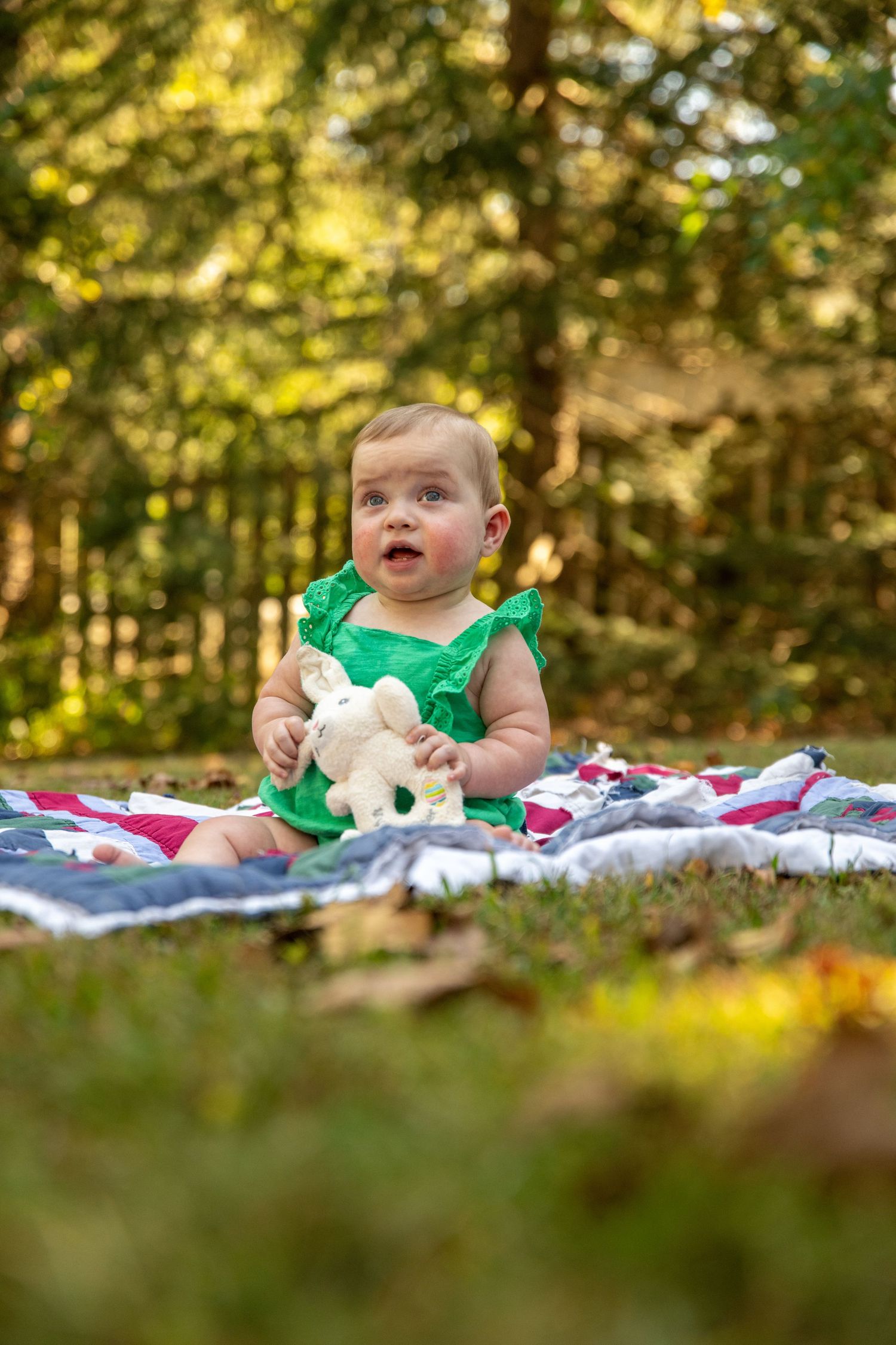 Baby in emerald dress playing on quilt in dappled sunlight under autumn trees.