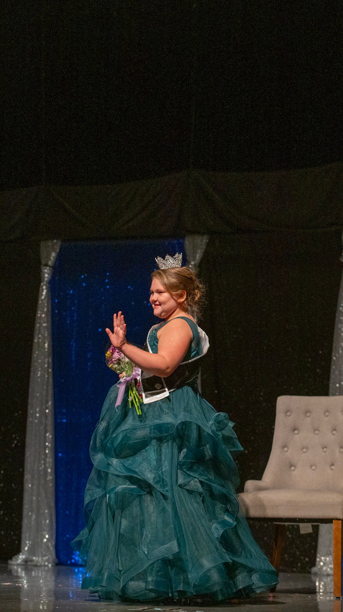 Pageant contestant in teal ballgown holds flowers while waving on stage with silver backdrop.