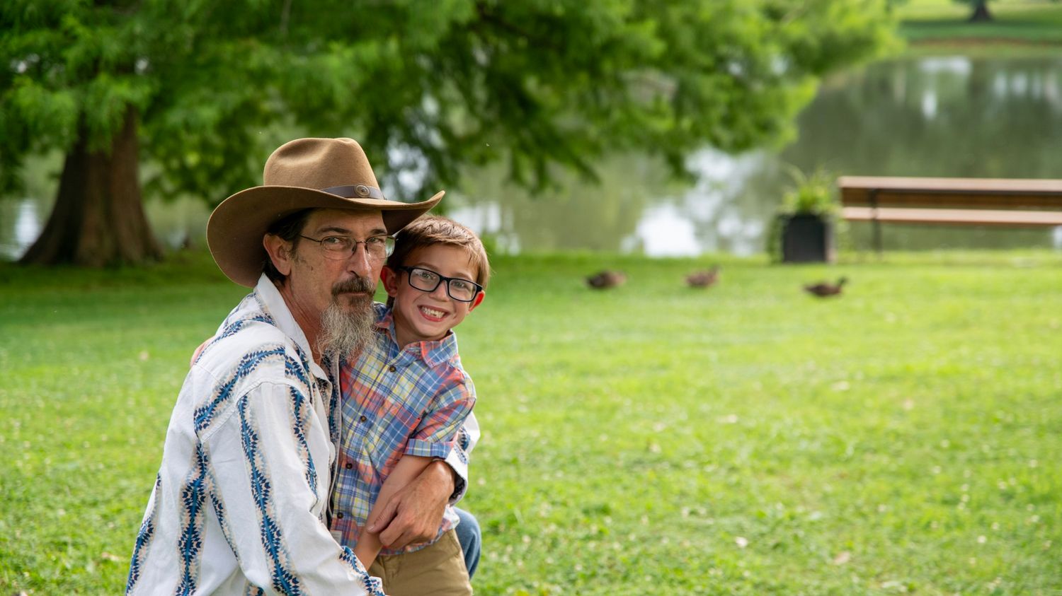 Two people share a happy moment together on grass with trees and pond in background.