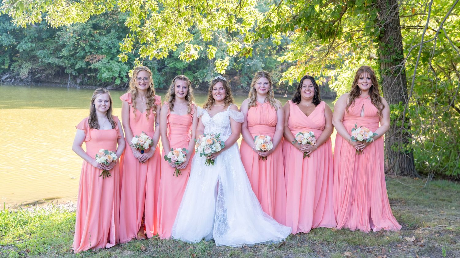 Wedding party in coral and white dresses poses together on grassy lawn.