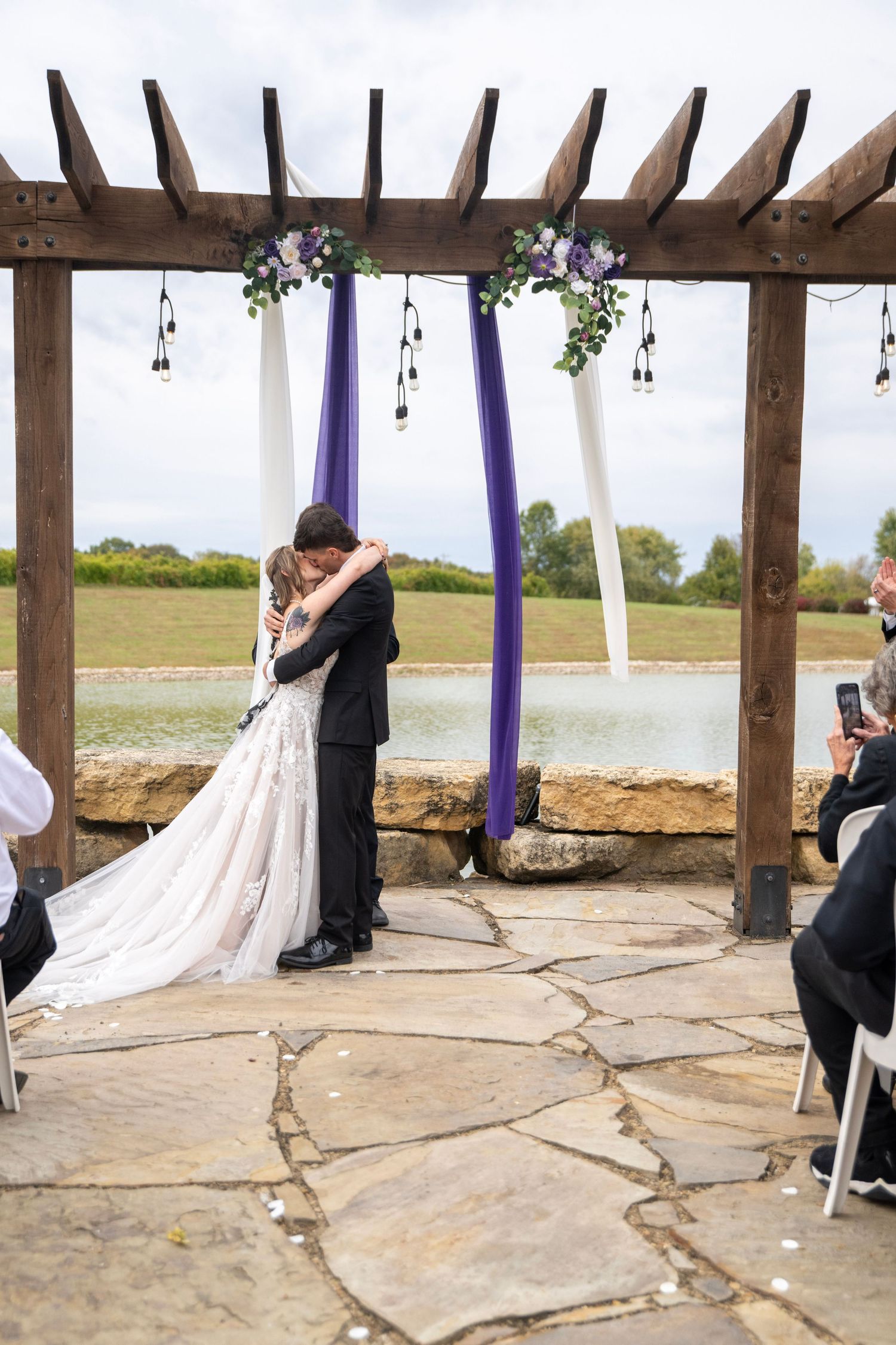 Wedding ceremony under wooden pergola decorated with white drapes and purple accents.