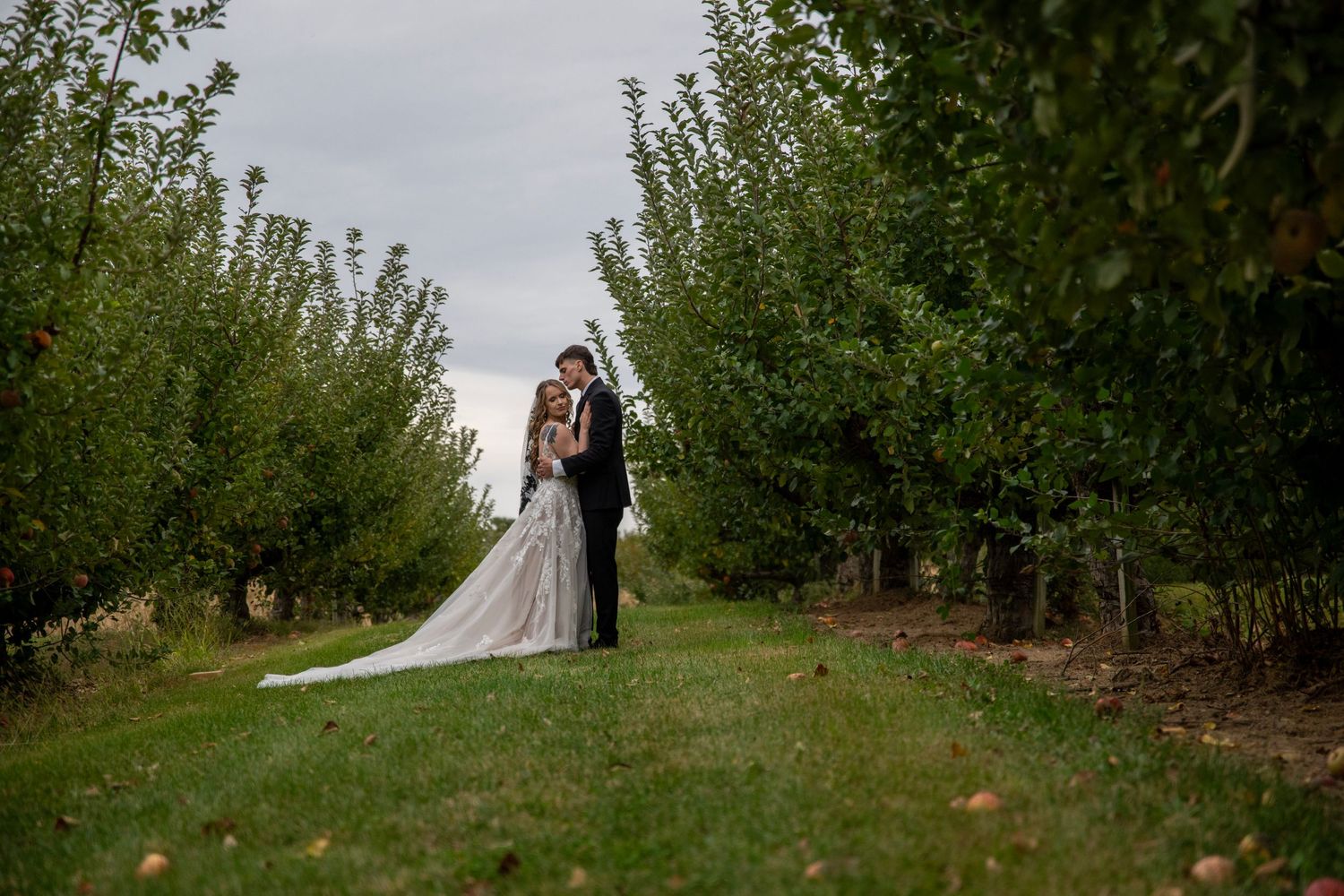 Wedding couple embraces in serene orchard setting with dramatic cloudy sky.