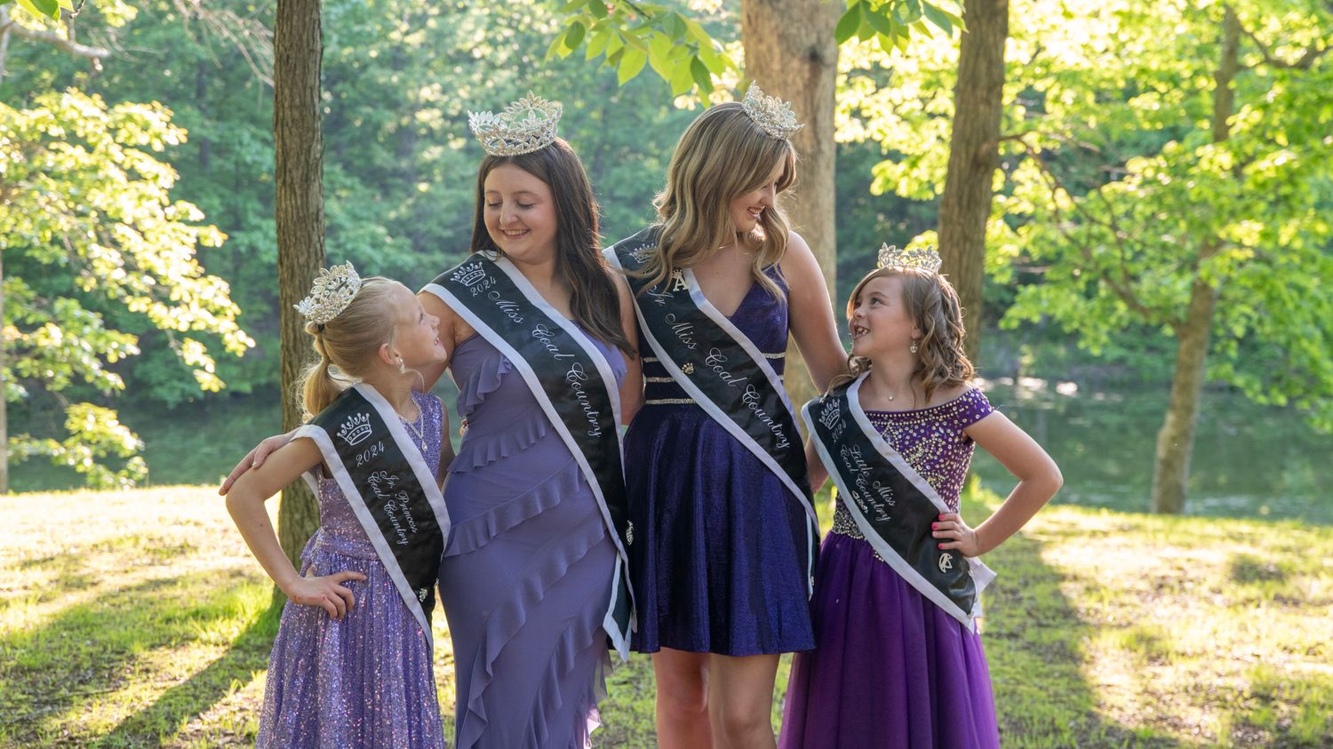 Pageant contestants in purple dresses pose together in a sunlit wooded area.