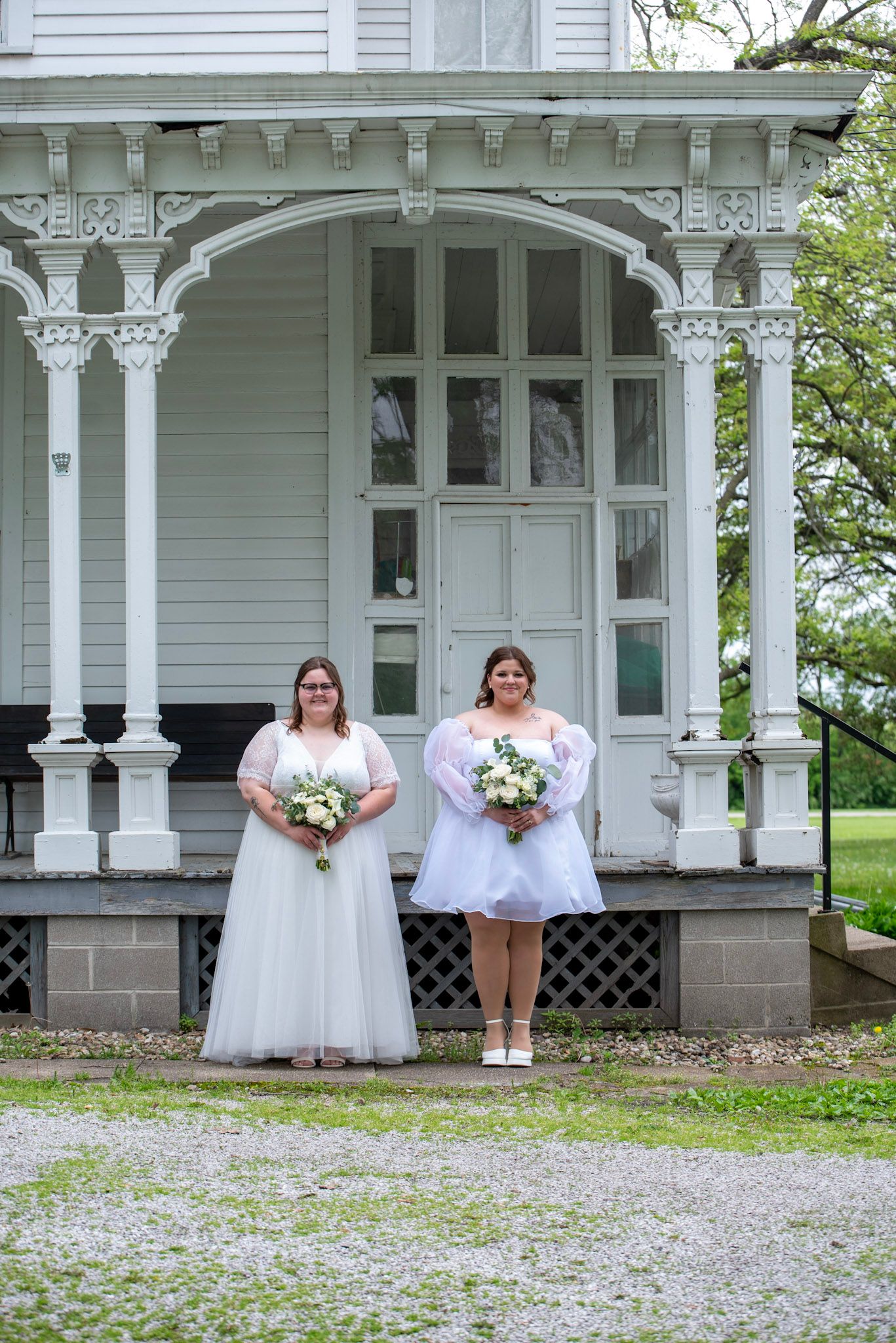 Two people in white dresses pose in front of historic white house with ornate porch details.