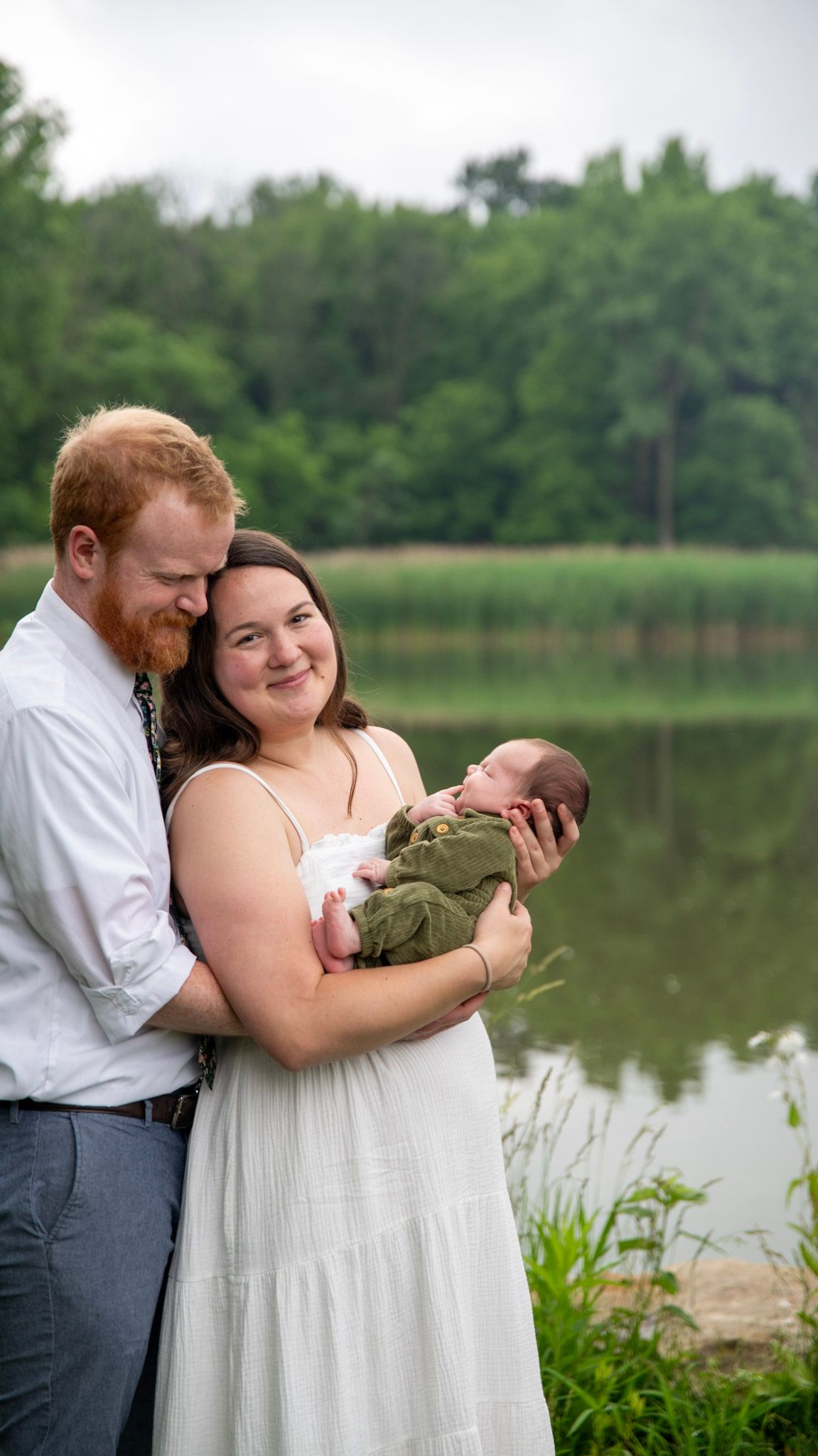 A couple shares tender moments with their baby by a serene lake surrounded by greenery.