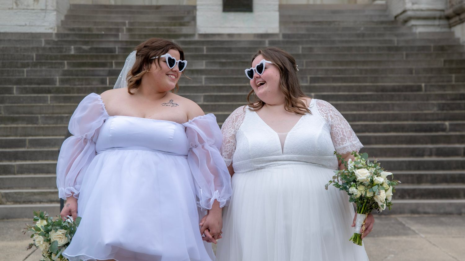 Two people in white dresses and sunglasses pose together against stone architecture.