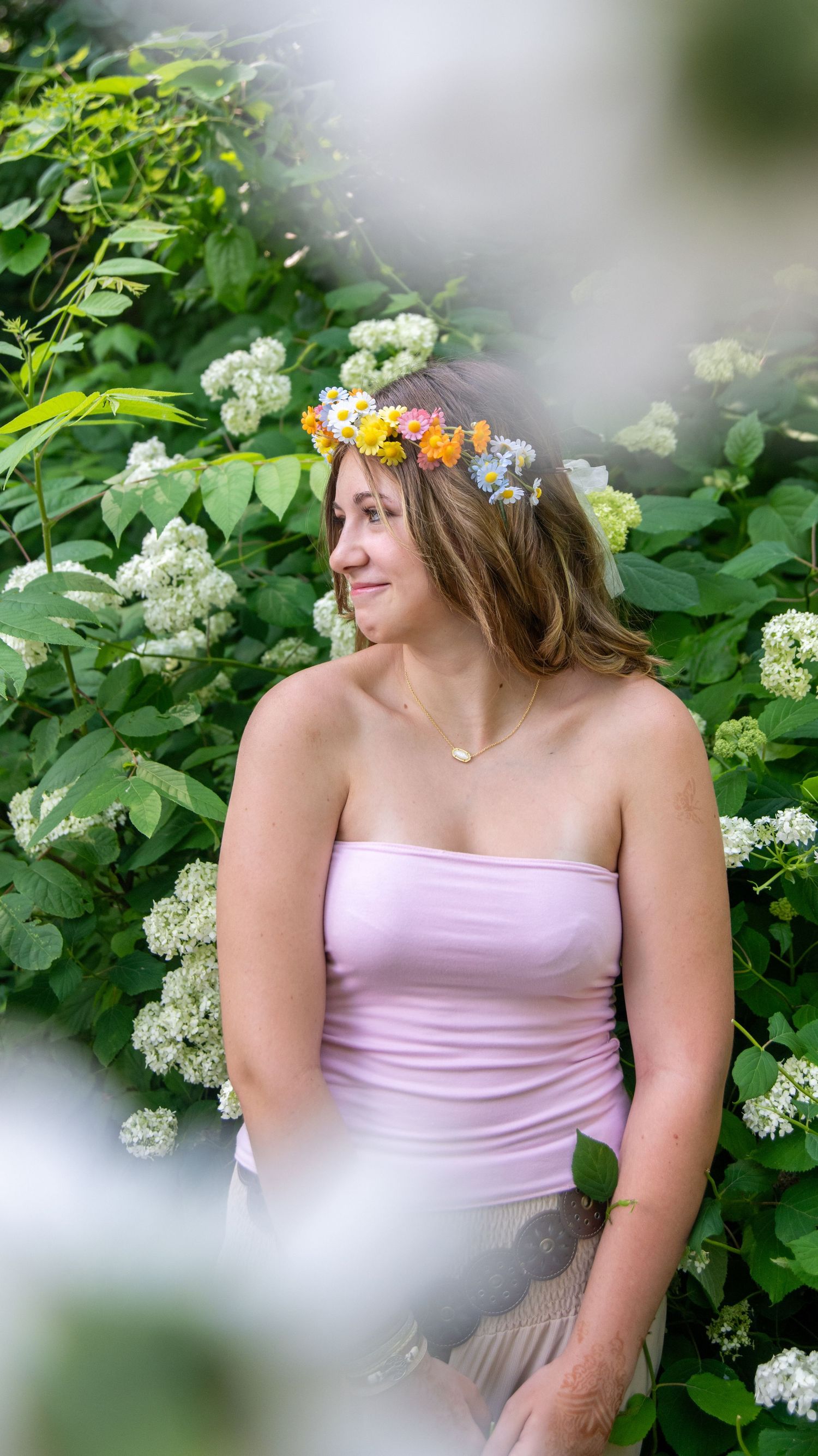 Portrait in soft pink dress with floral crown against white flowering bushes in garden setting.