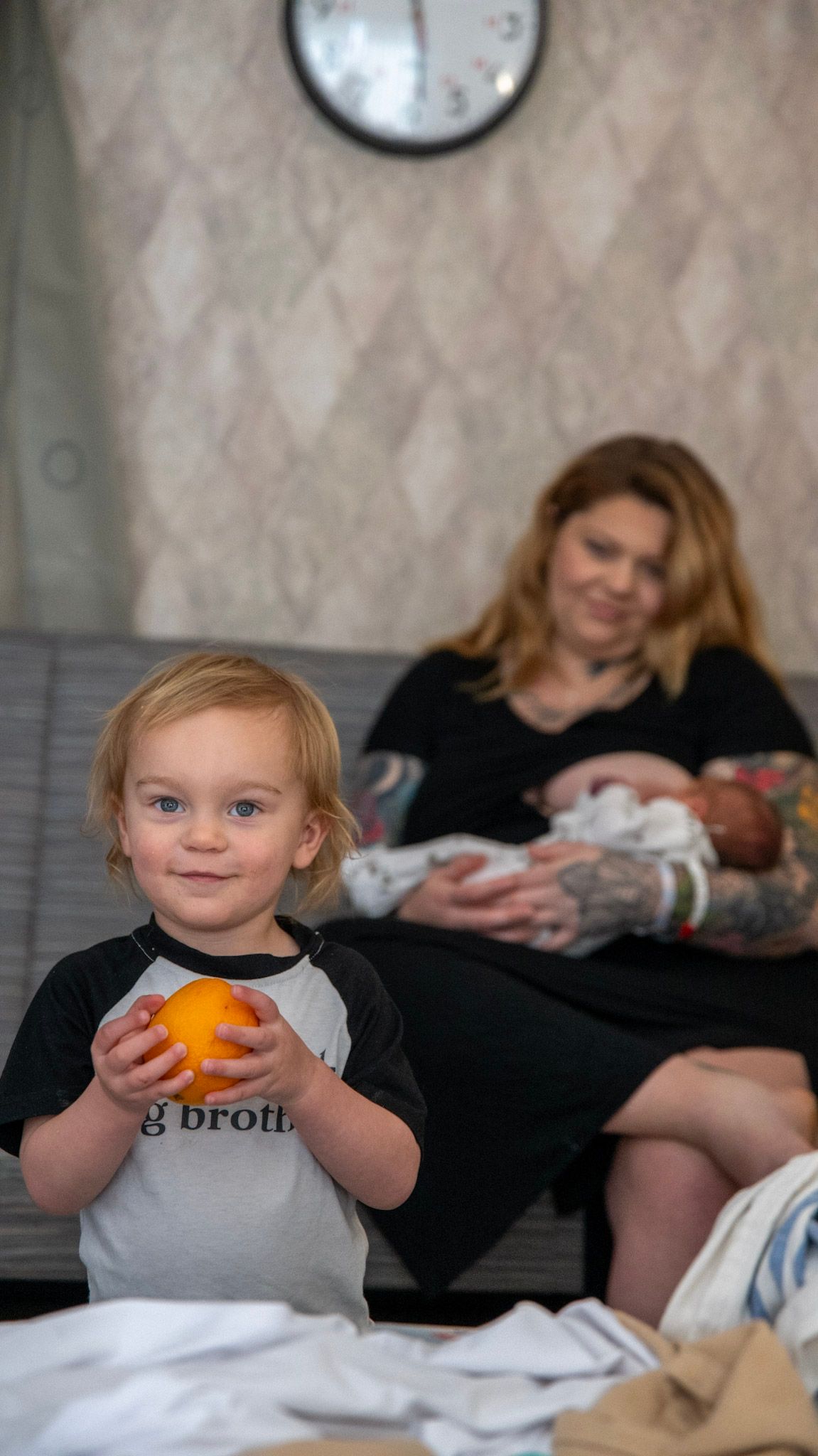 A toddler holds an orange while adults interact with a baby in the background.
