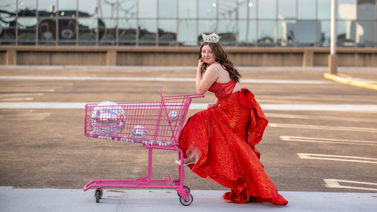 A creative fashion shoot combining a flowing red gown with pink shopping carts on a concrete surface.