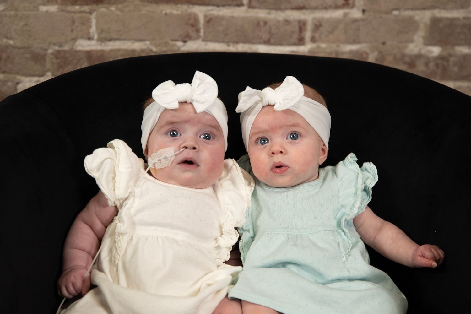 Twin babies in coordinating pastel dresses and white bow headbands pose on dark furniture.