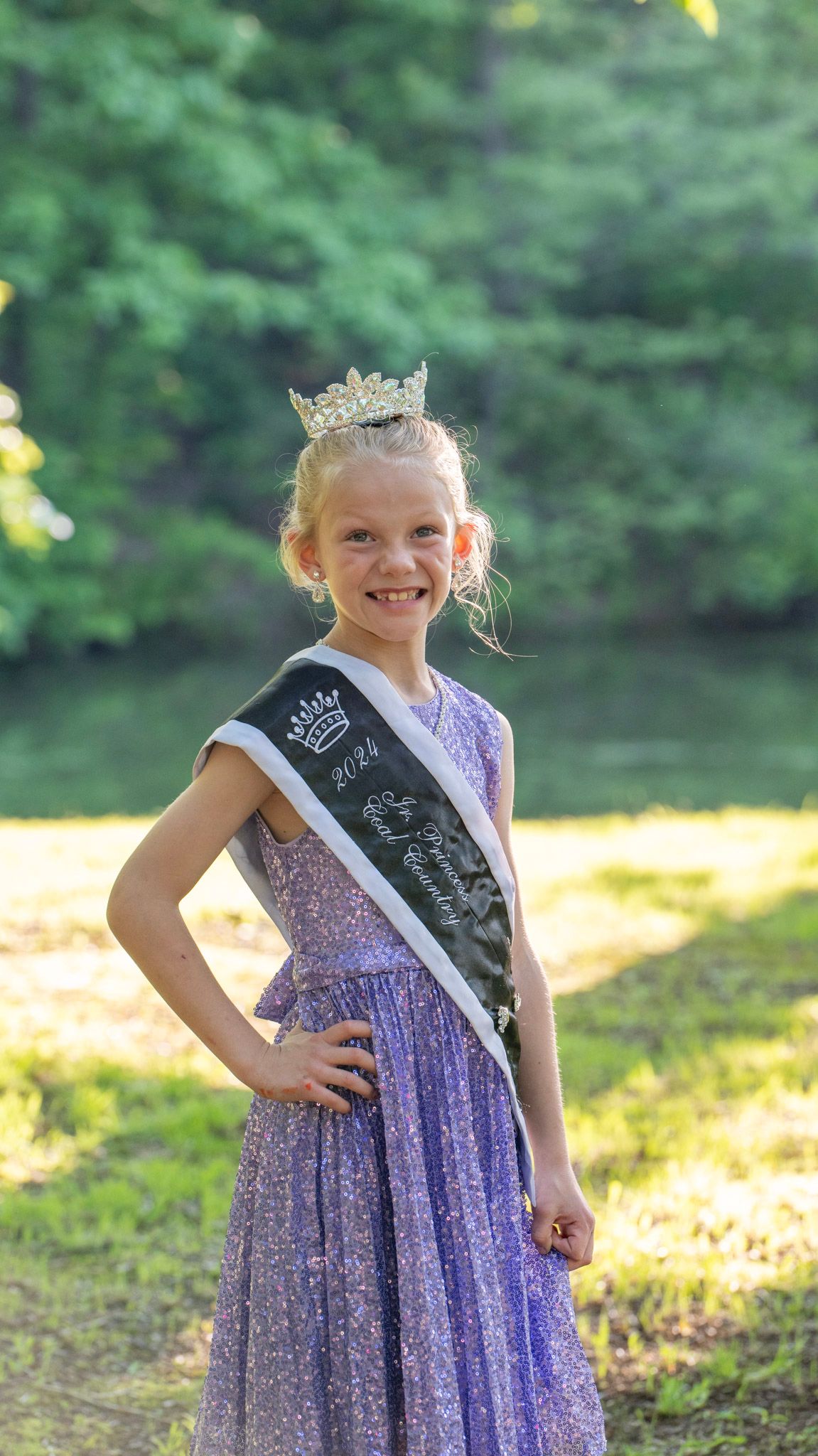 Young contestant in purple dress and crown poses outdoors in natural lighting.