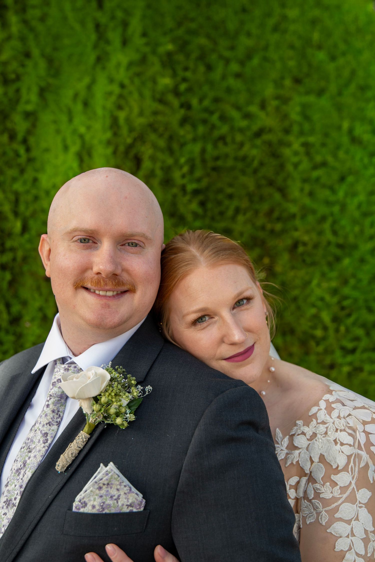 Wedding portrait against lush green hedge background with formal attire and floral boutonniere.