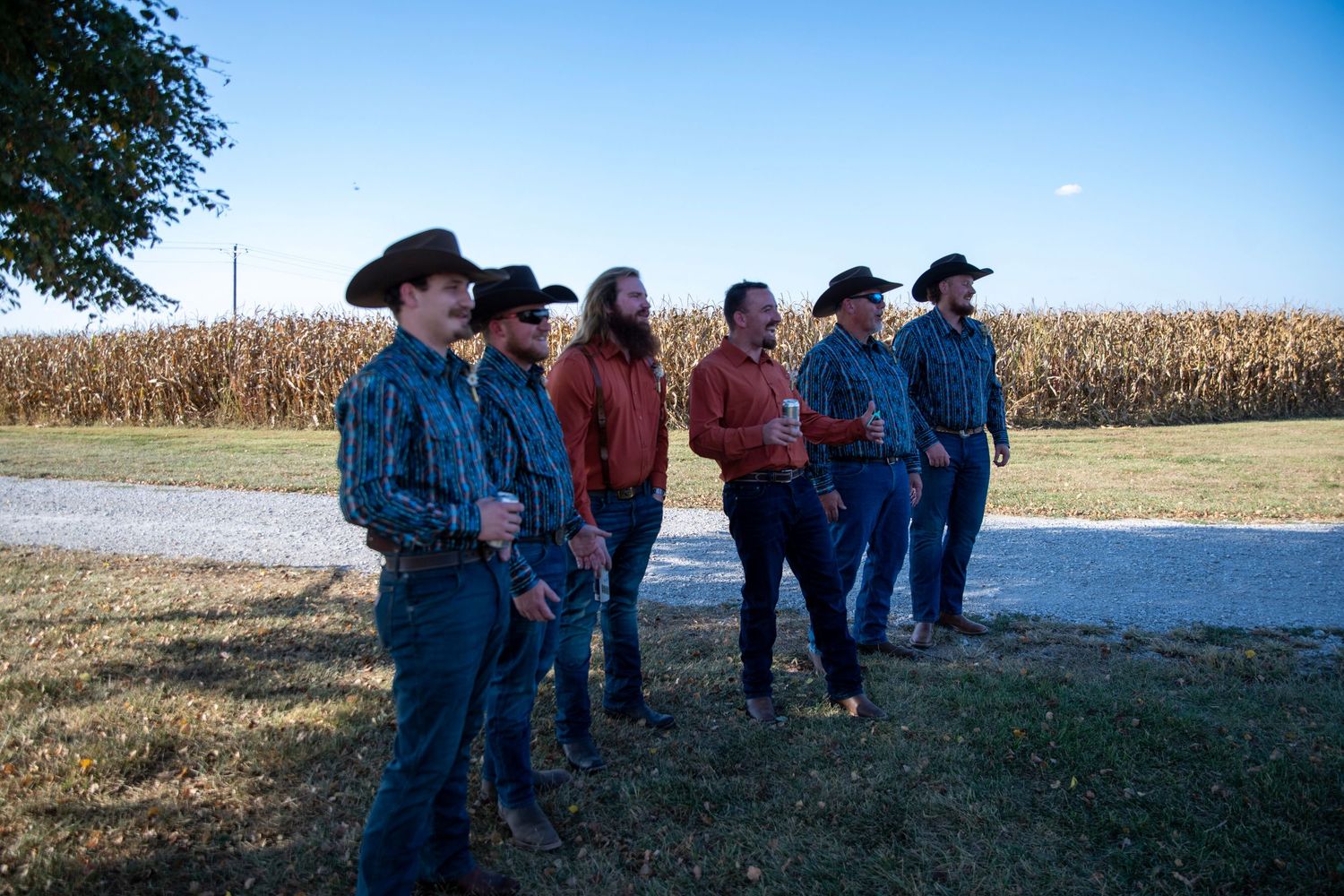 People in western attire gather outdoors near a cornfield on a sunny day.