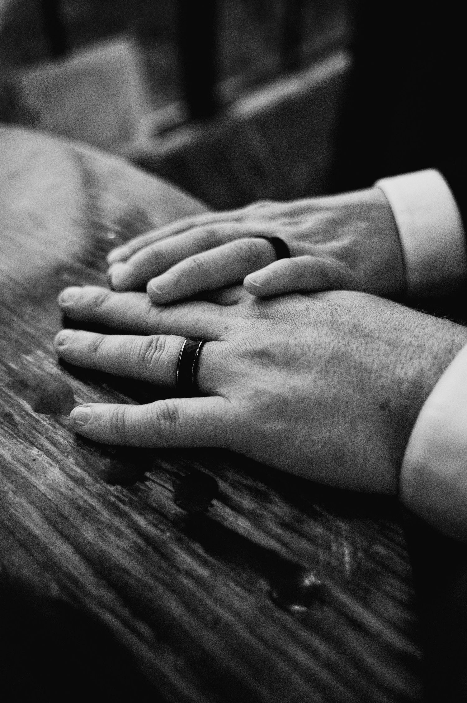 Black and white close-up of hands with wedding rings resting on a textured surface.
