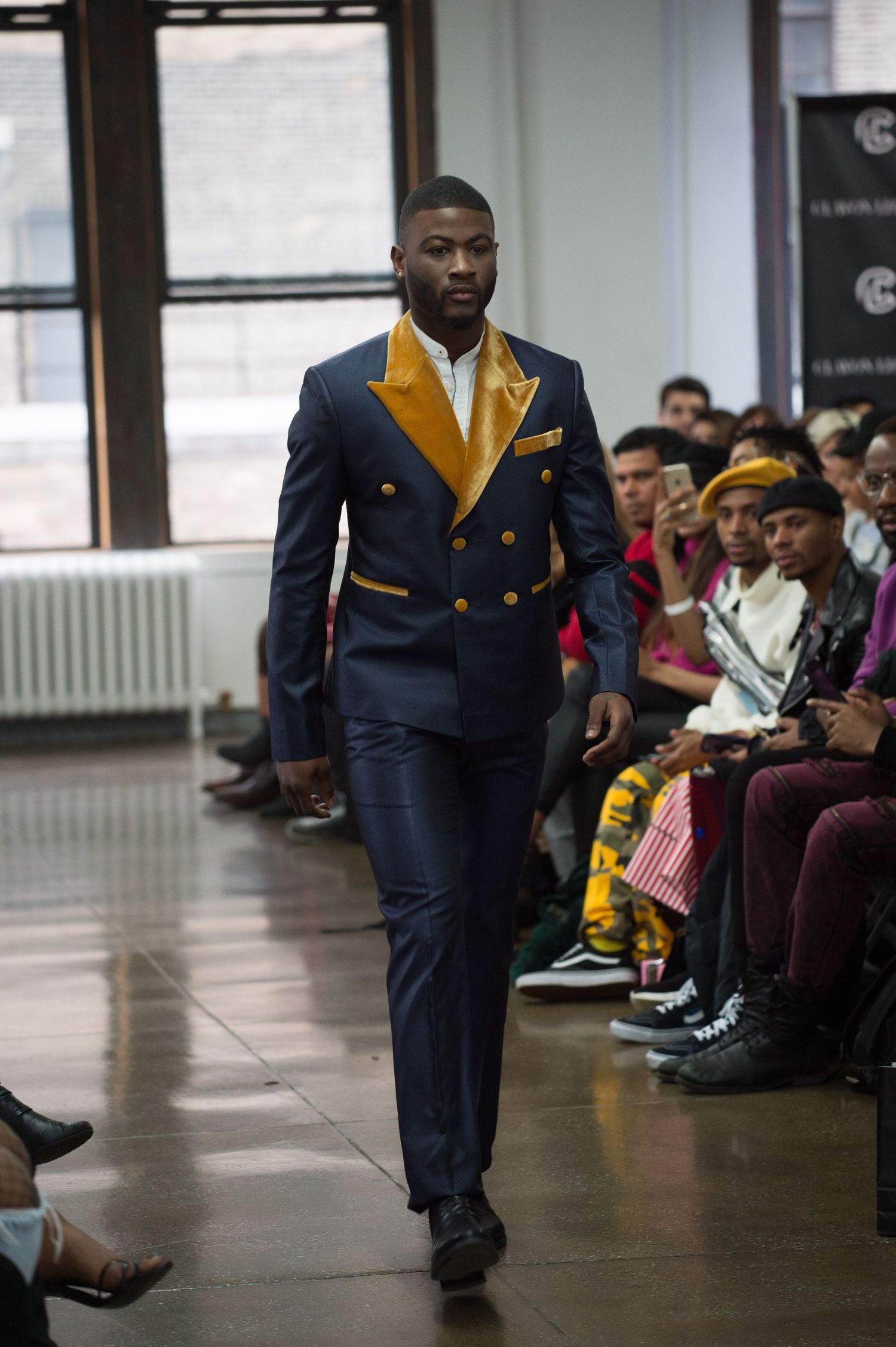 Model walks runway in navy and yellow formal wear during indoor fashion show.