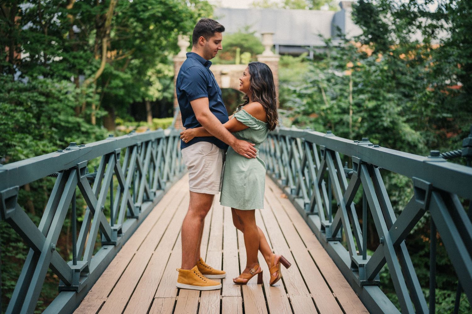 Couple embraces on wooden footbridge surrounded by lush green foliage during outdoor engagement photo session.