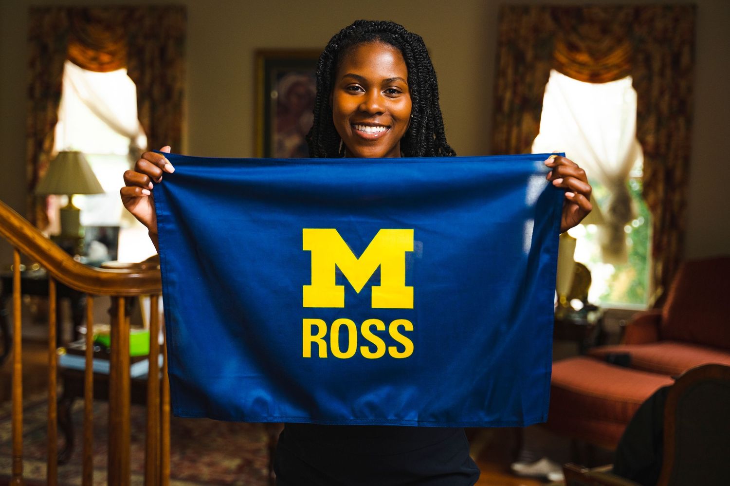 Person holding up a blue and yellow University of Michigan banner in a home setting.