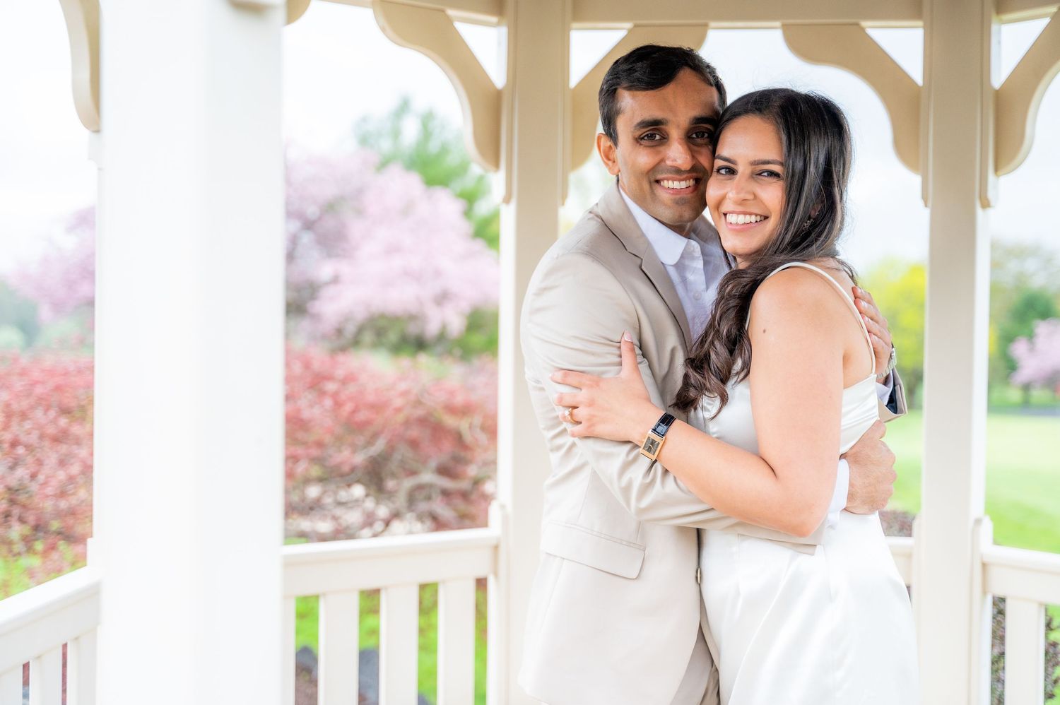 A romantic couple embraces on a white gazebo porch surrounded by blooming pink spring trees.