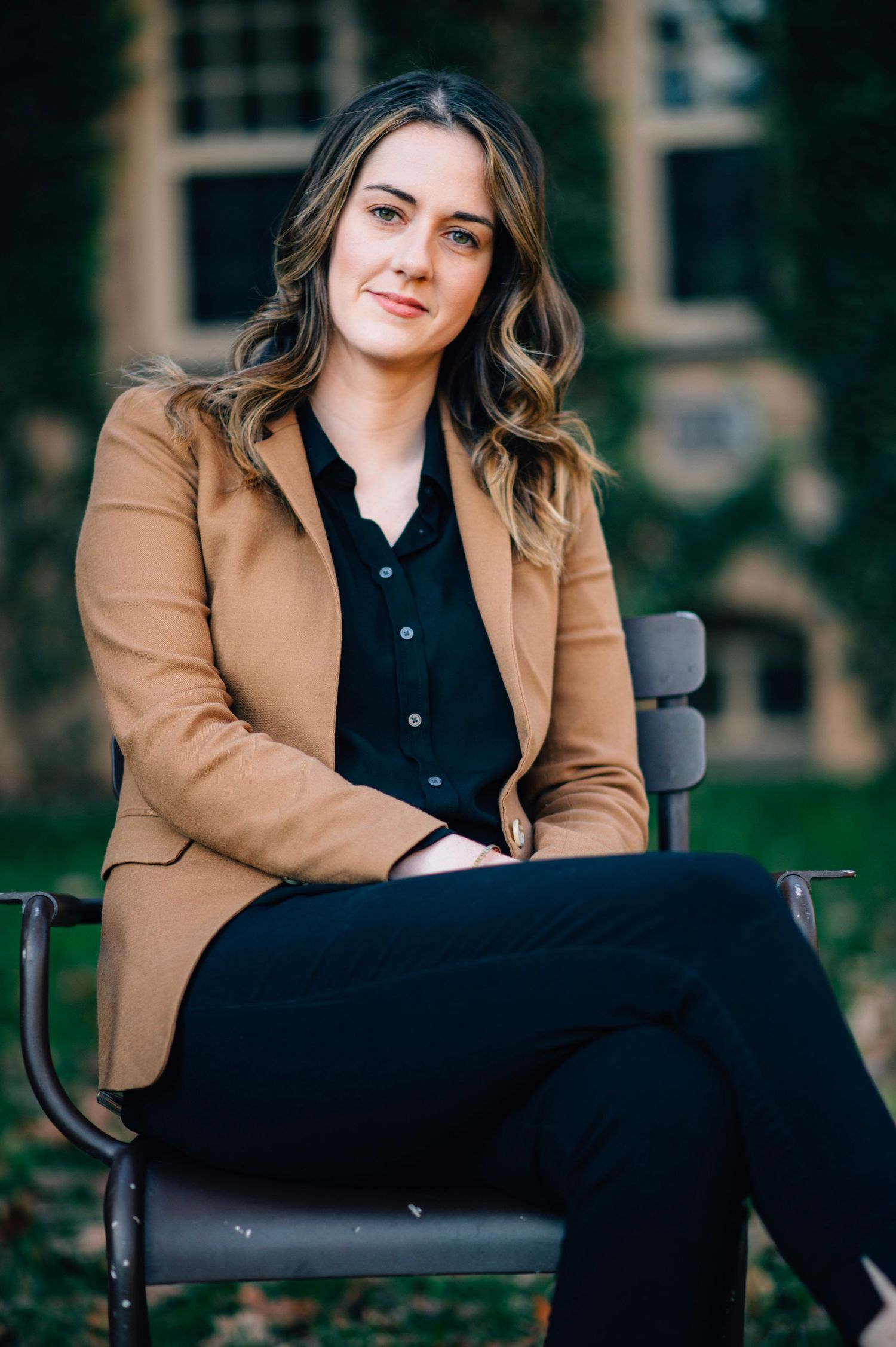 Professional portrait on a bench wearing a tan blazer and black outfit.