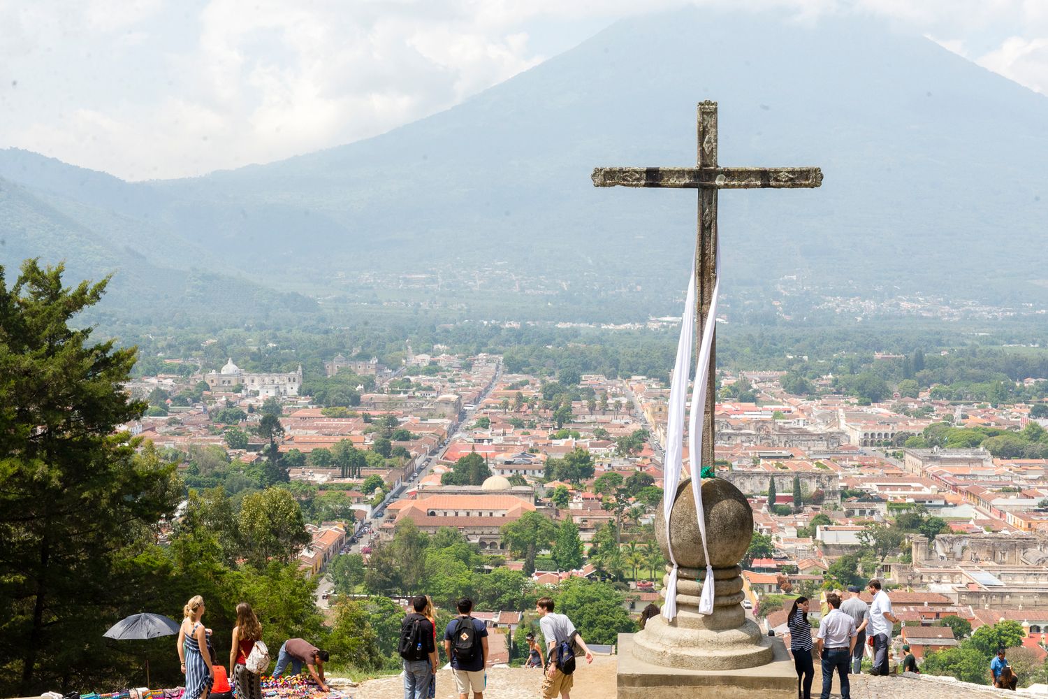 Tourists gather around a hilltop cross overlooking the scenic cityscape of Antigua Guatemala with mountains in the background.