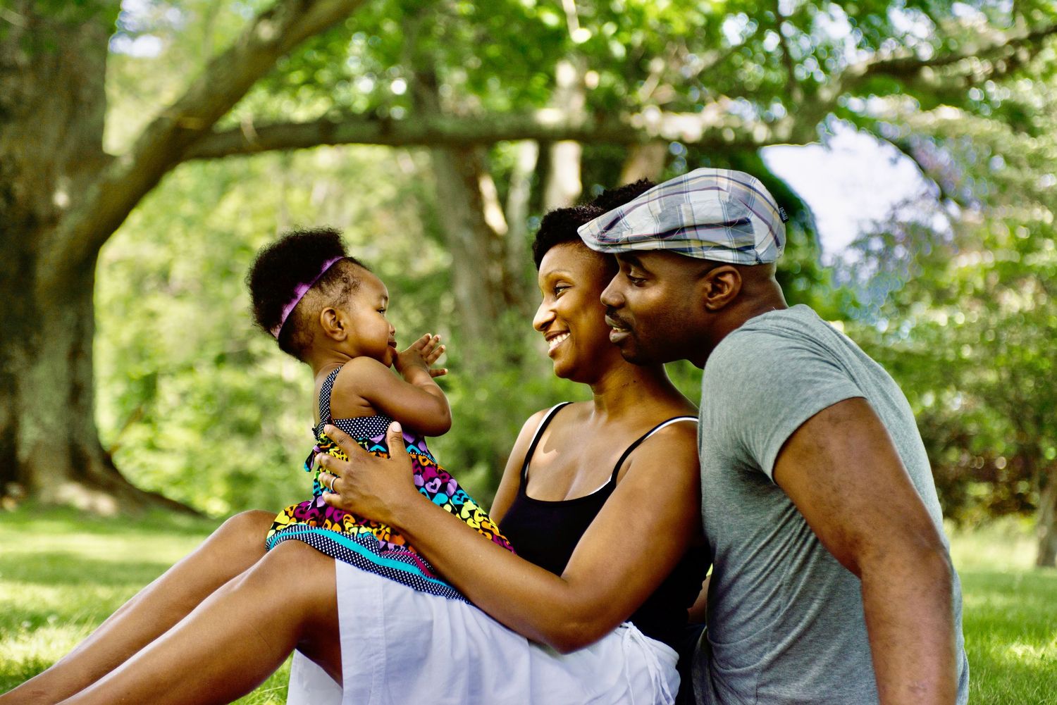 A couple shares a tender moment outdoors in a park setting with lush green trees in the background.
