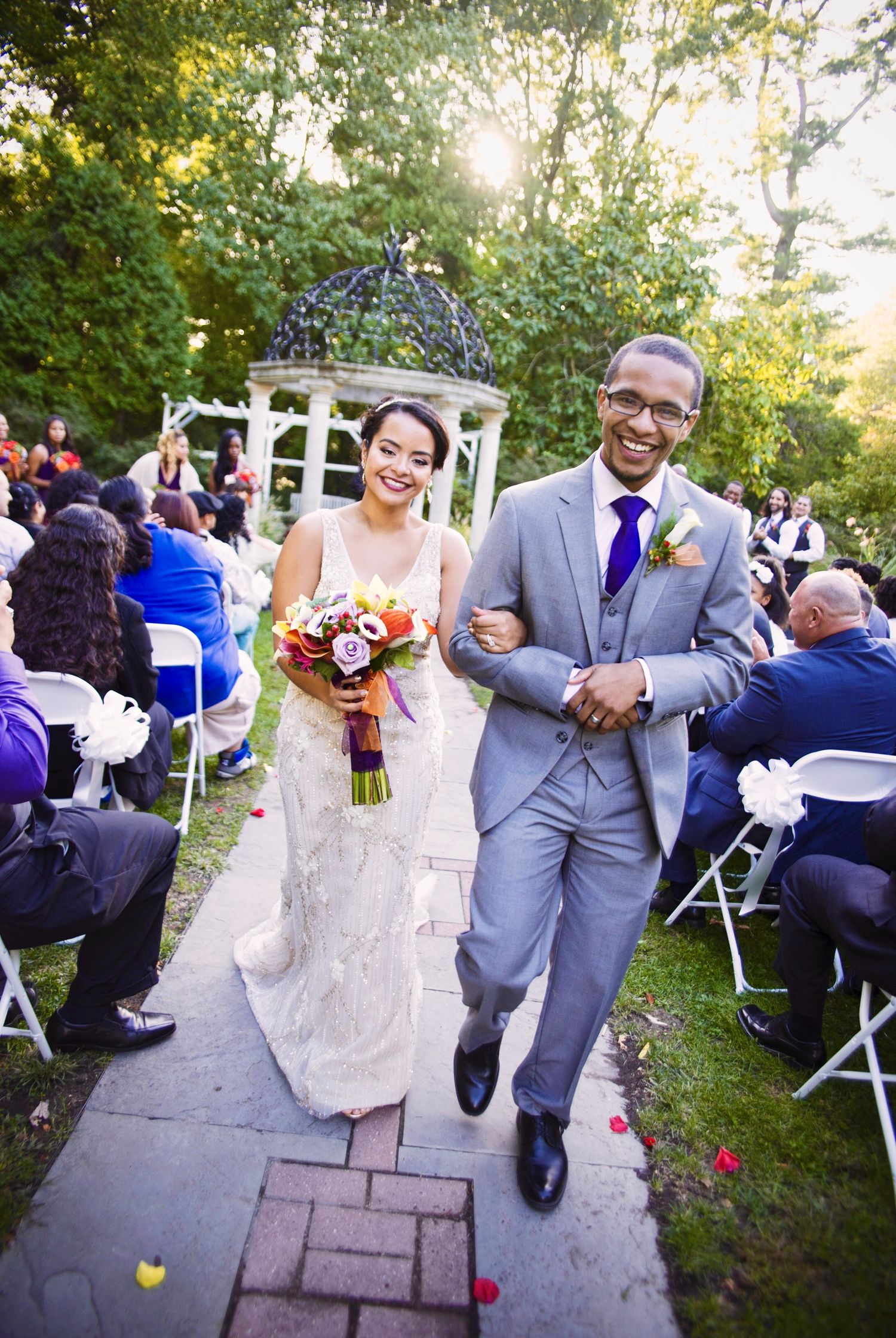 Newlyweds walk down the aisle after their outdoor garden ceremony as guests shower them with colorful flower petals.