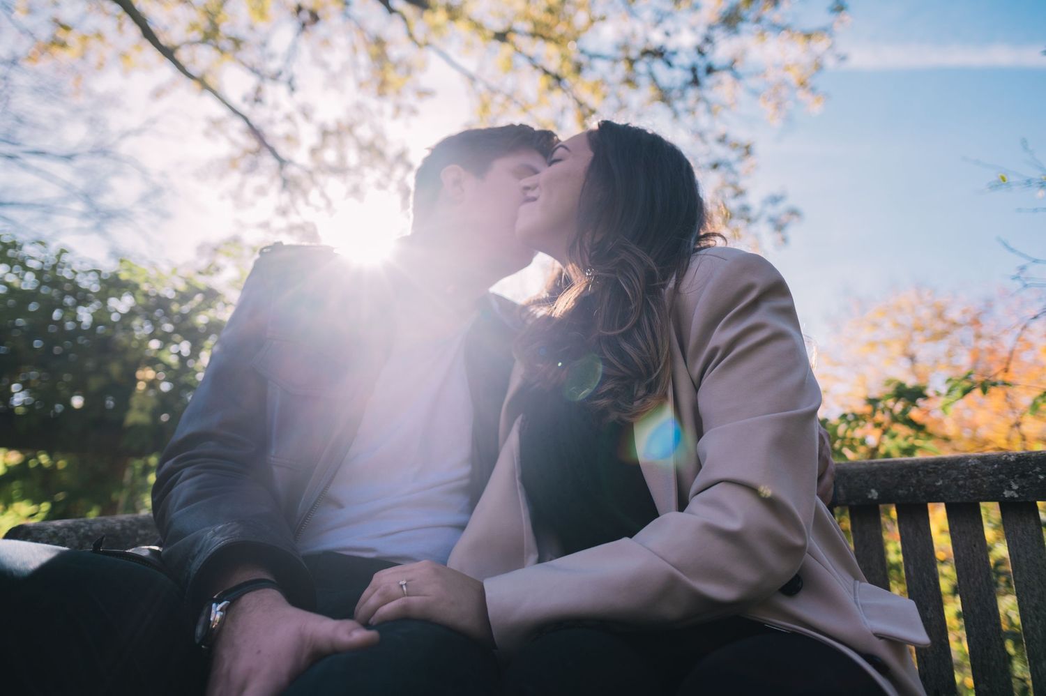 Two people share a tender moment on a wooden bench under autumn trees with sunlight streaming through the branches.