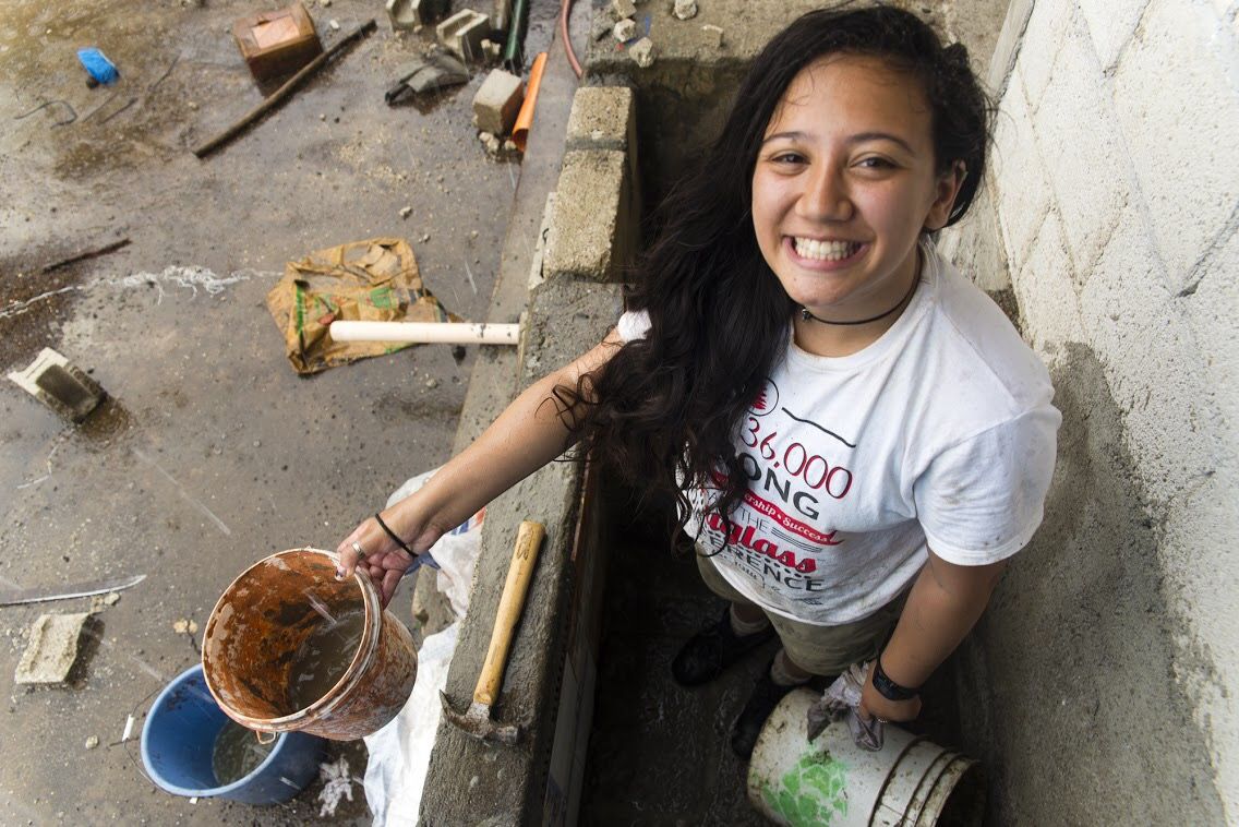 Someone in a white t-shirt smiles while working on plumbing repairs in a narrow space.